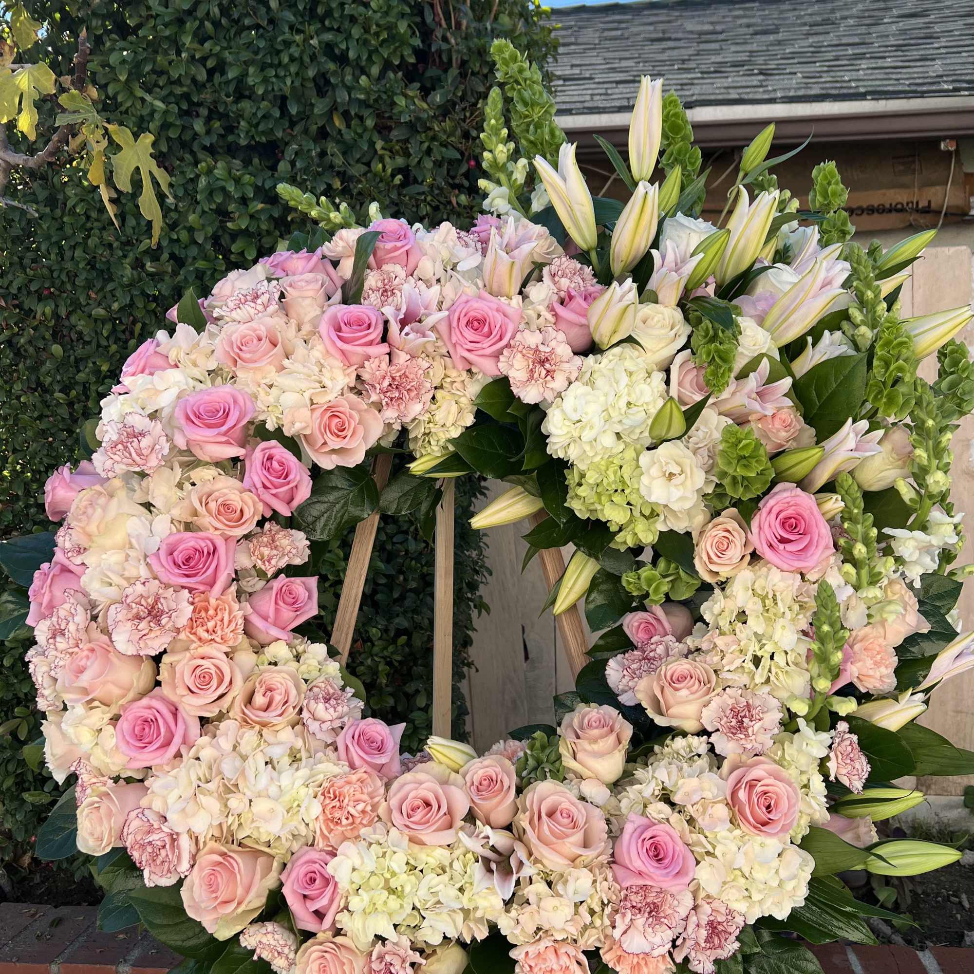 Large standing wreath of pink roses, white hydrangeas, carnations, and lilies on a wooden easel.