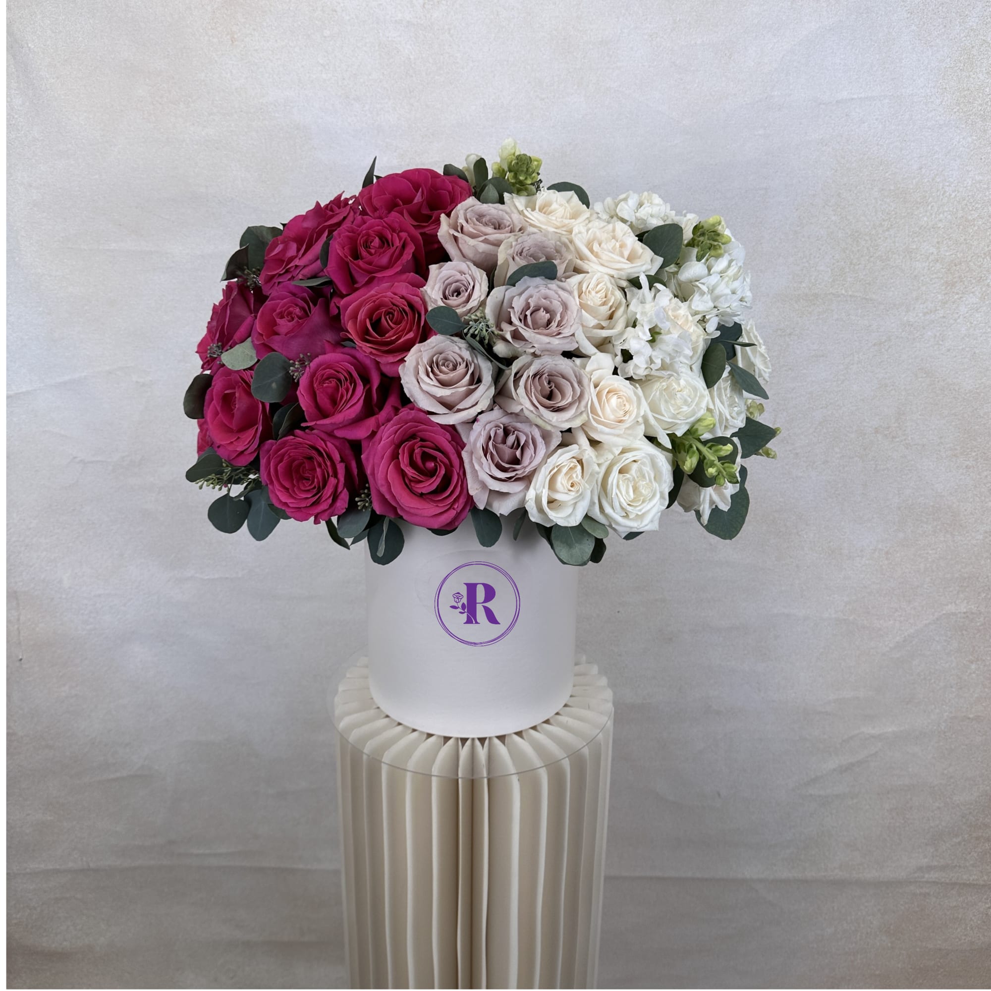 Round hatbox arrangement of pink, lavender, and white roses displayed on a cream pedestal stand