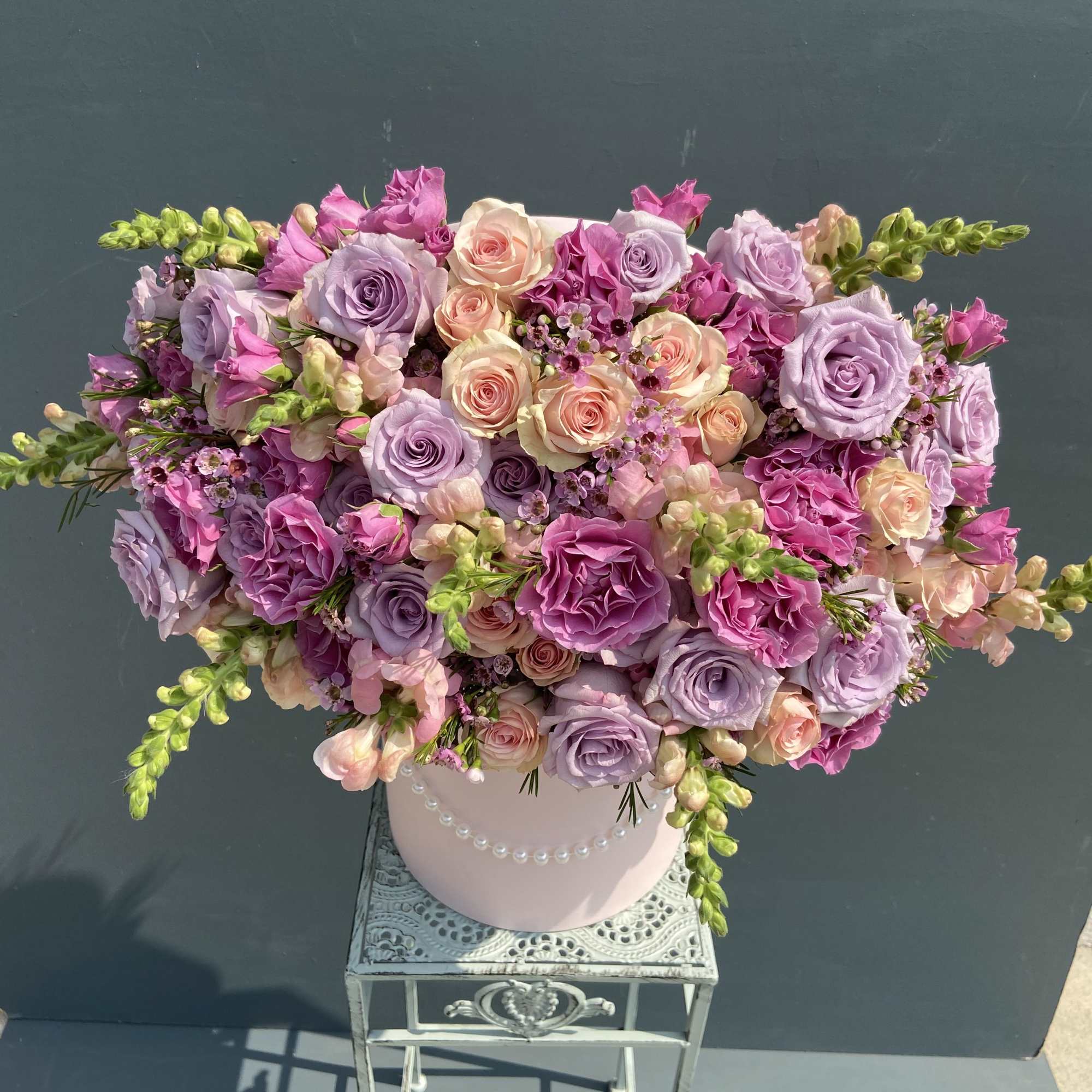 Round pink hatbox filled with pink, lavender and peach roses with snapdragons on a metal stand.