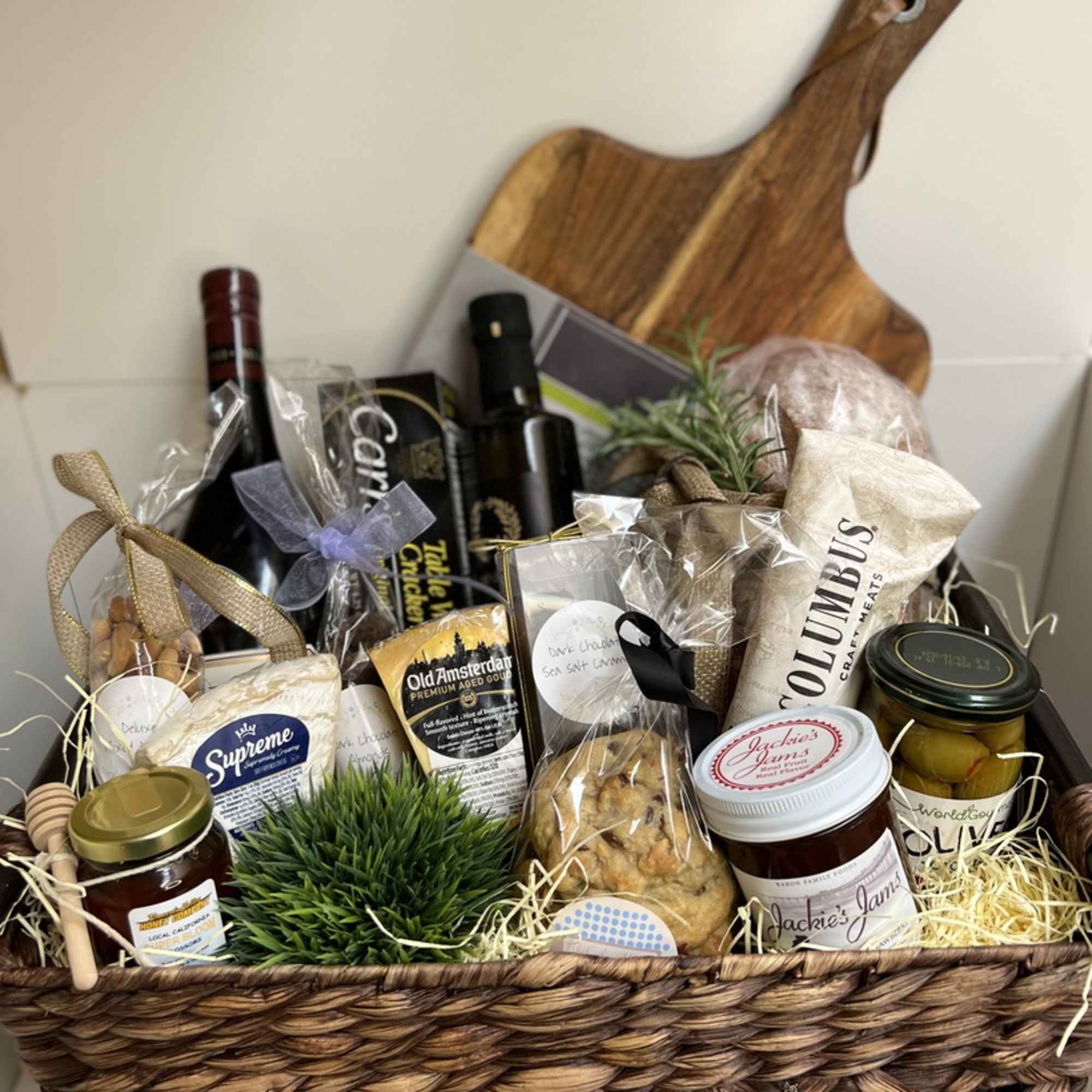 Gourmet gift basket with wine, cheeses, jars, baked goods, and a wooden cutting board.