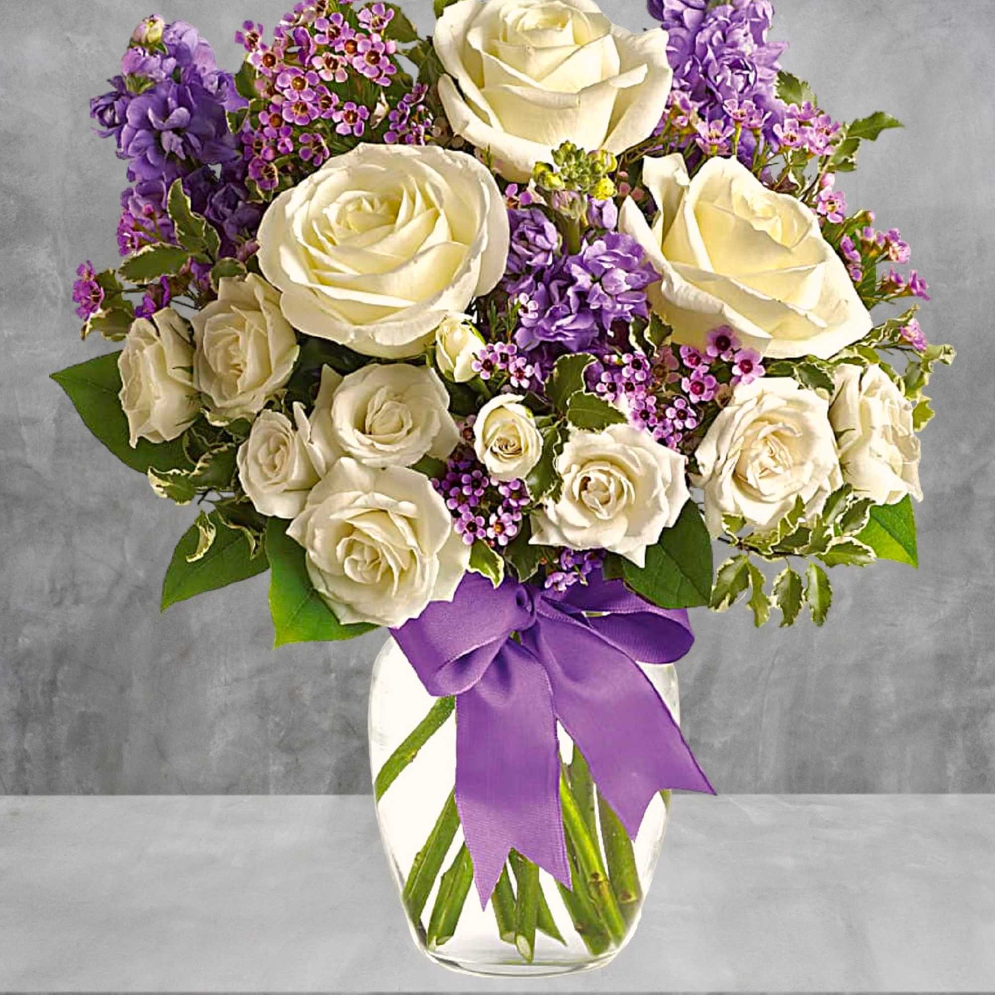 White roses and purple flowers in a clear vase with a purple ribbon