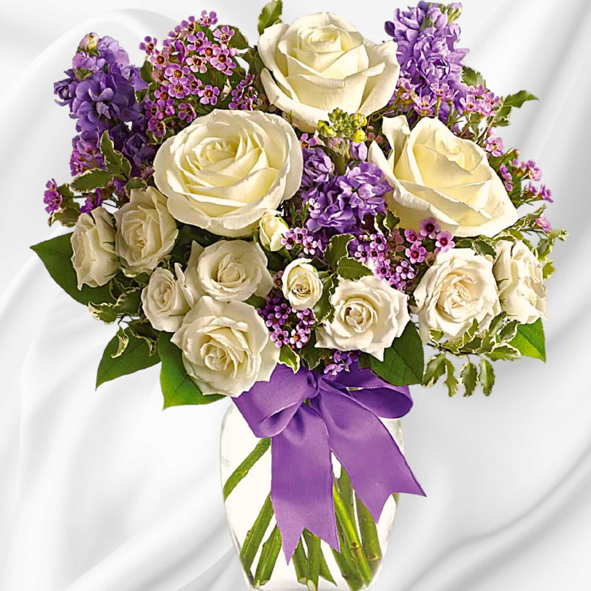 White roses and purple flowers arranged in a clear glass vase with a purple ribbon