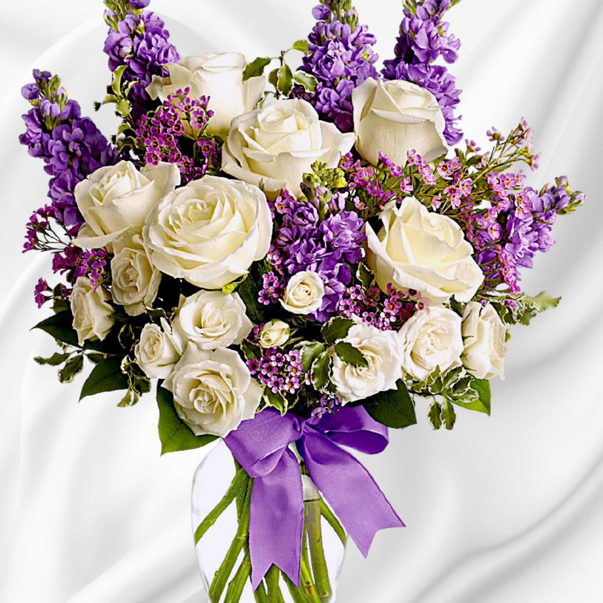 White roses and purple flowers in a clear glass vase with a purple ribbon