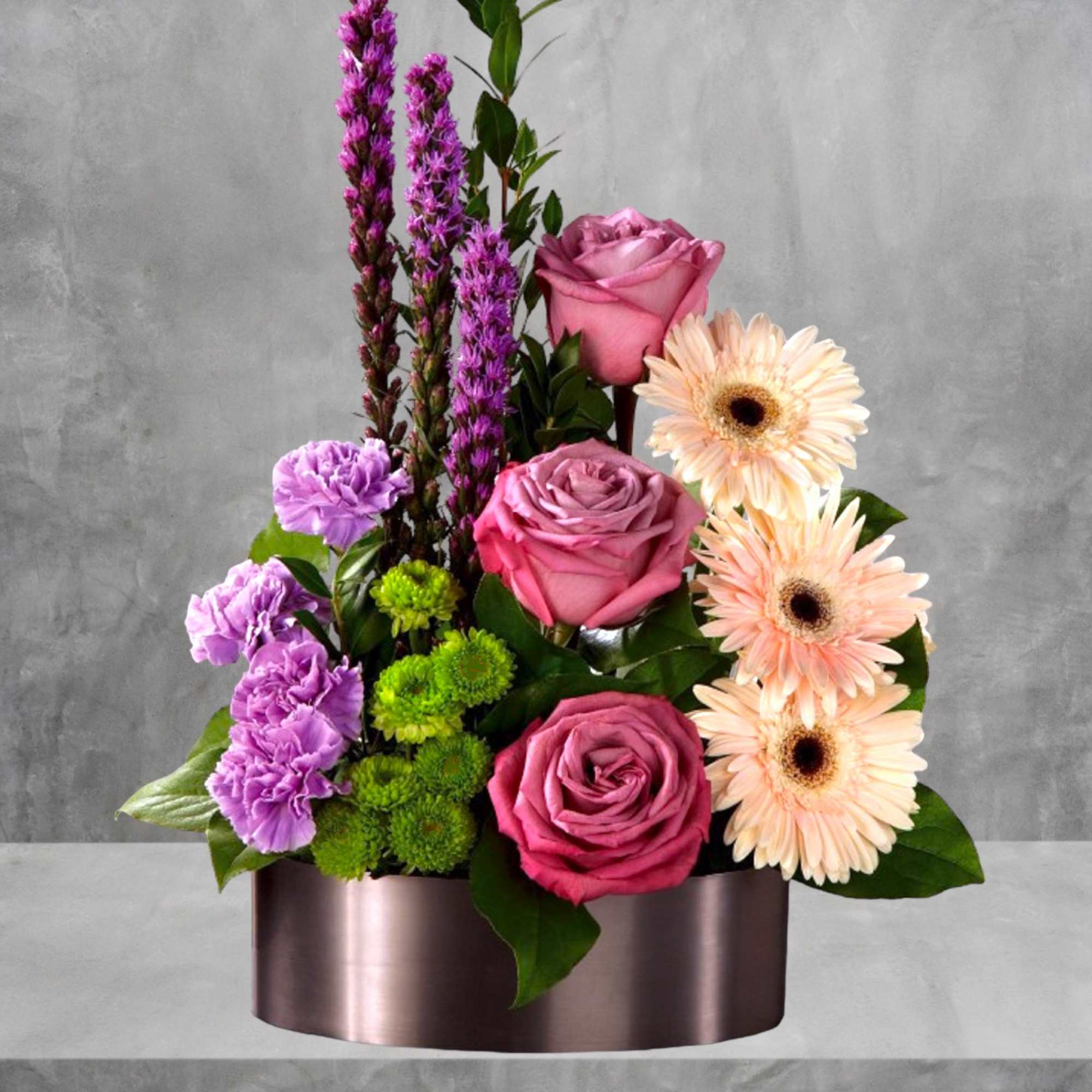 Pink roses, gerbera daisies, and purple blooms in a round metallic box