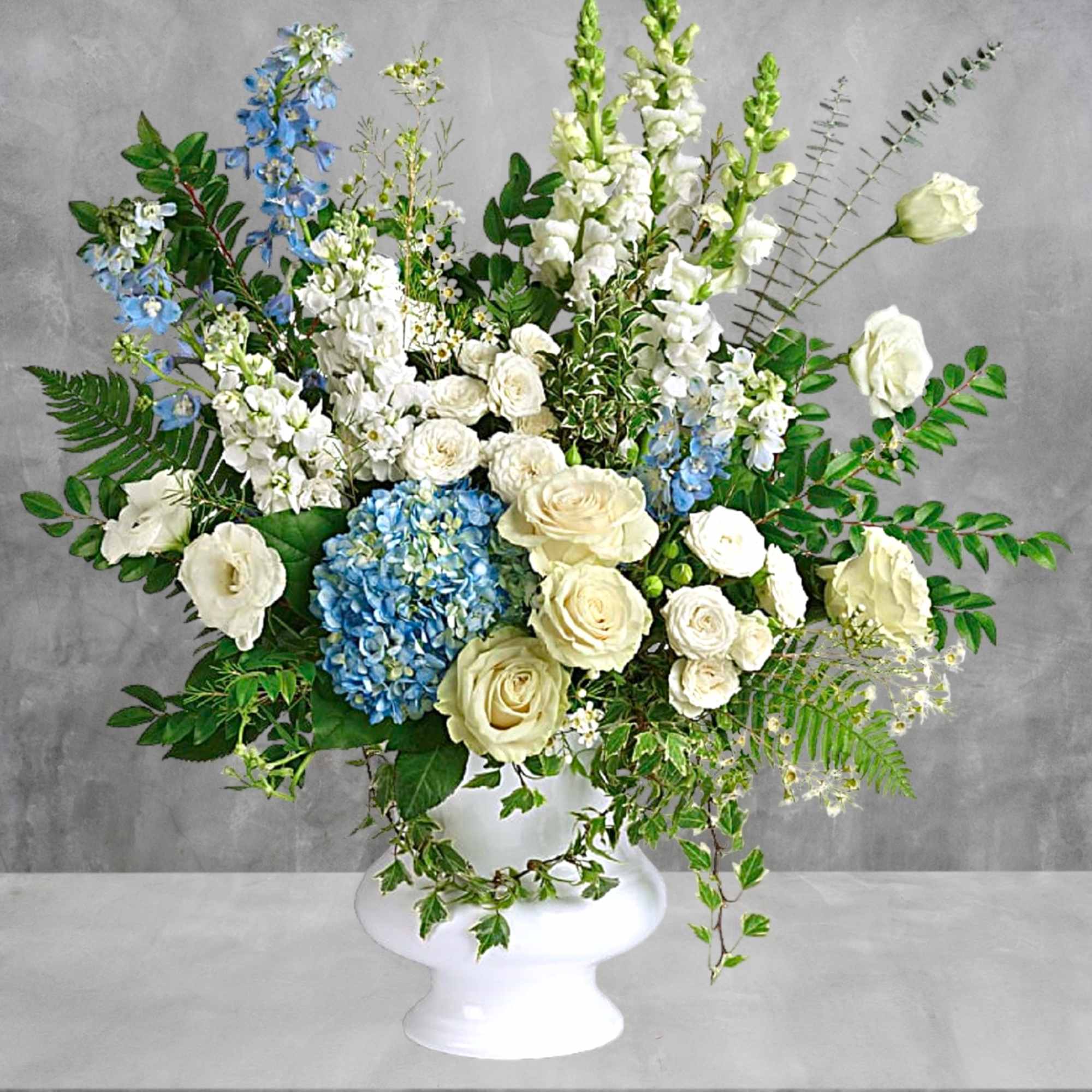 Large white vase arrangement with cream roses, blue hydrangea, and tall white blooms