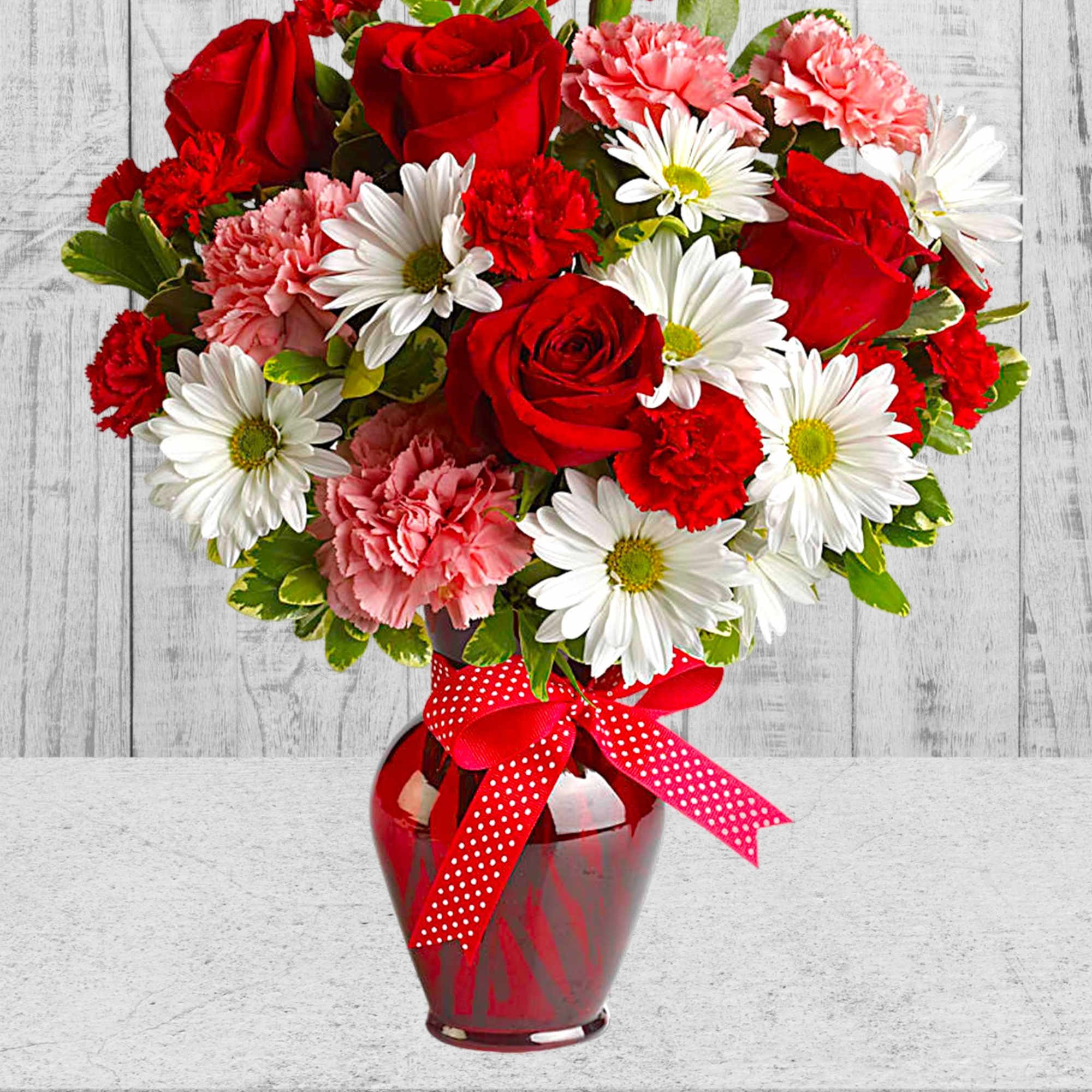 Red roses, white daisies, and pink carnations in a red vase with a polka-dot ribbon.
