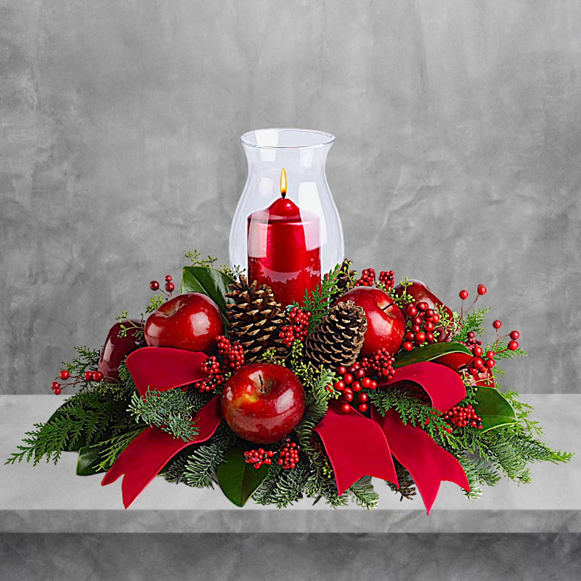 Red candle centerpiece with apples, pinecones, and ribbon in a glass hurricane