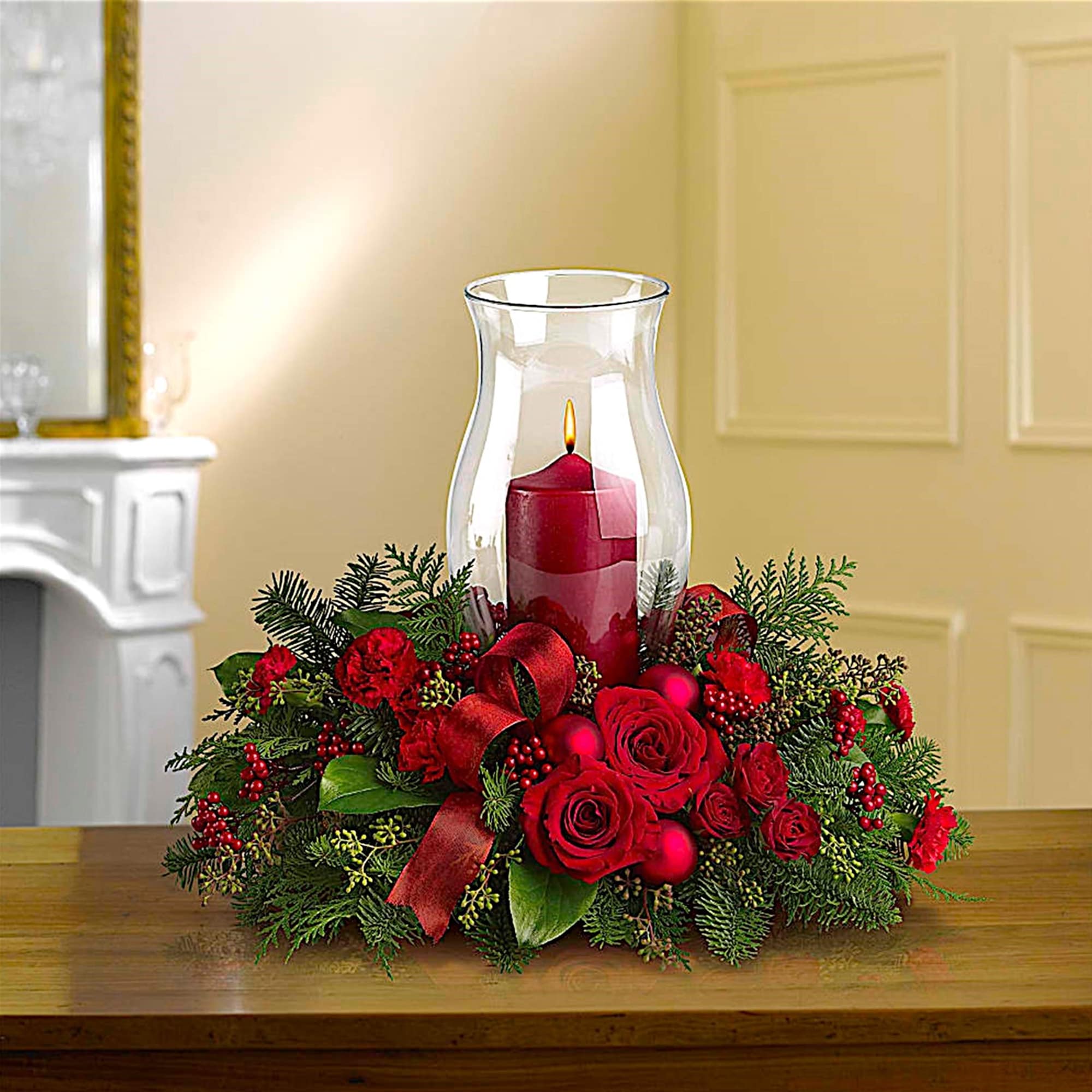 Red roses and greenery surround a lit candle in a glass hurricane vase.