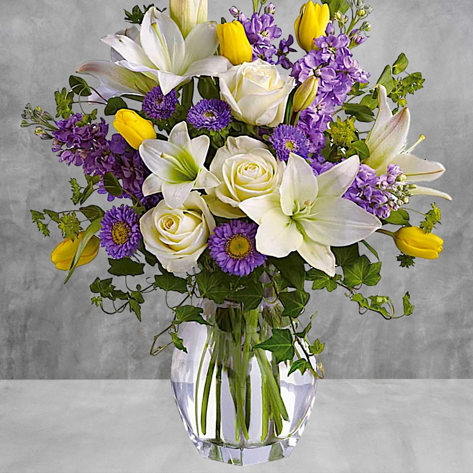 Bouquet of white lilies, roses, purple flowers, and yellow tulips in a clear glass vase