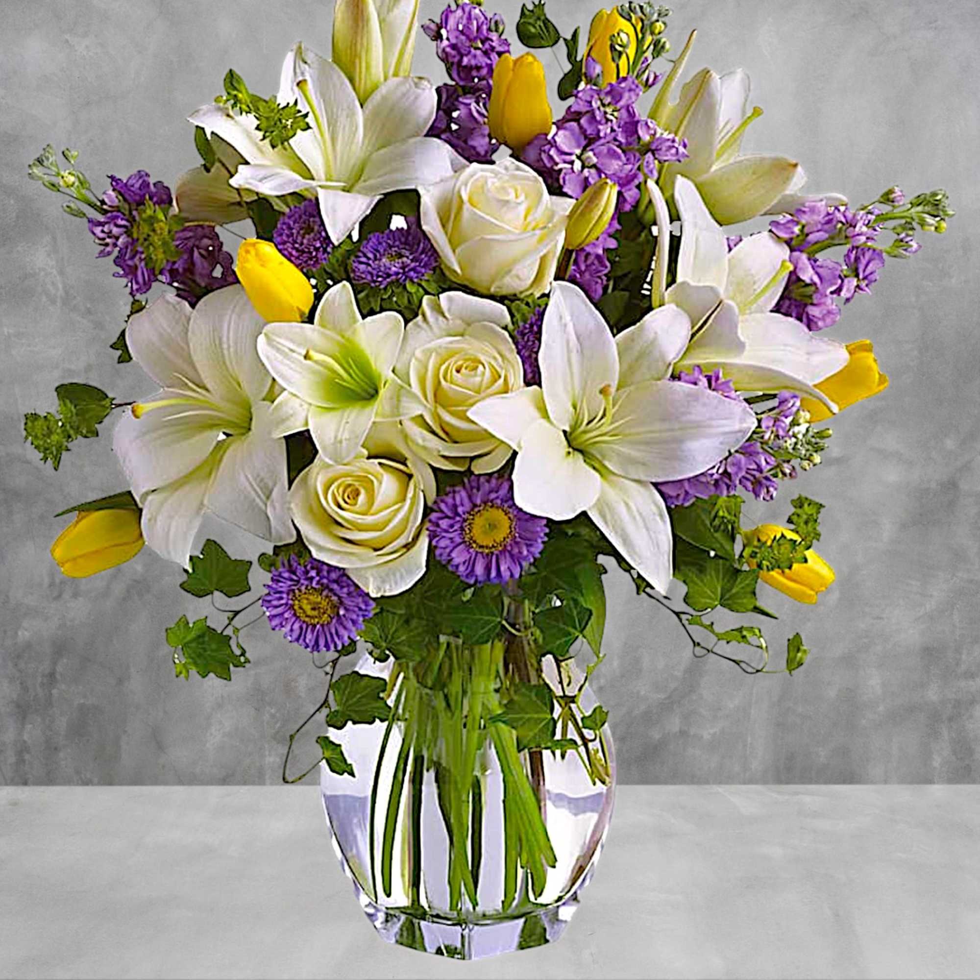 Bouquet of white lilies, yellow tulips, and purple flowers in a glass vase