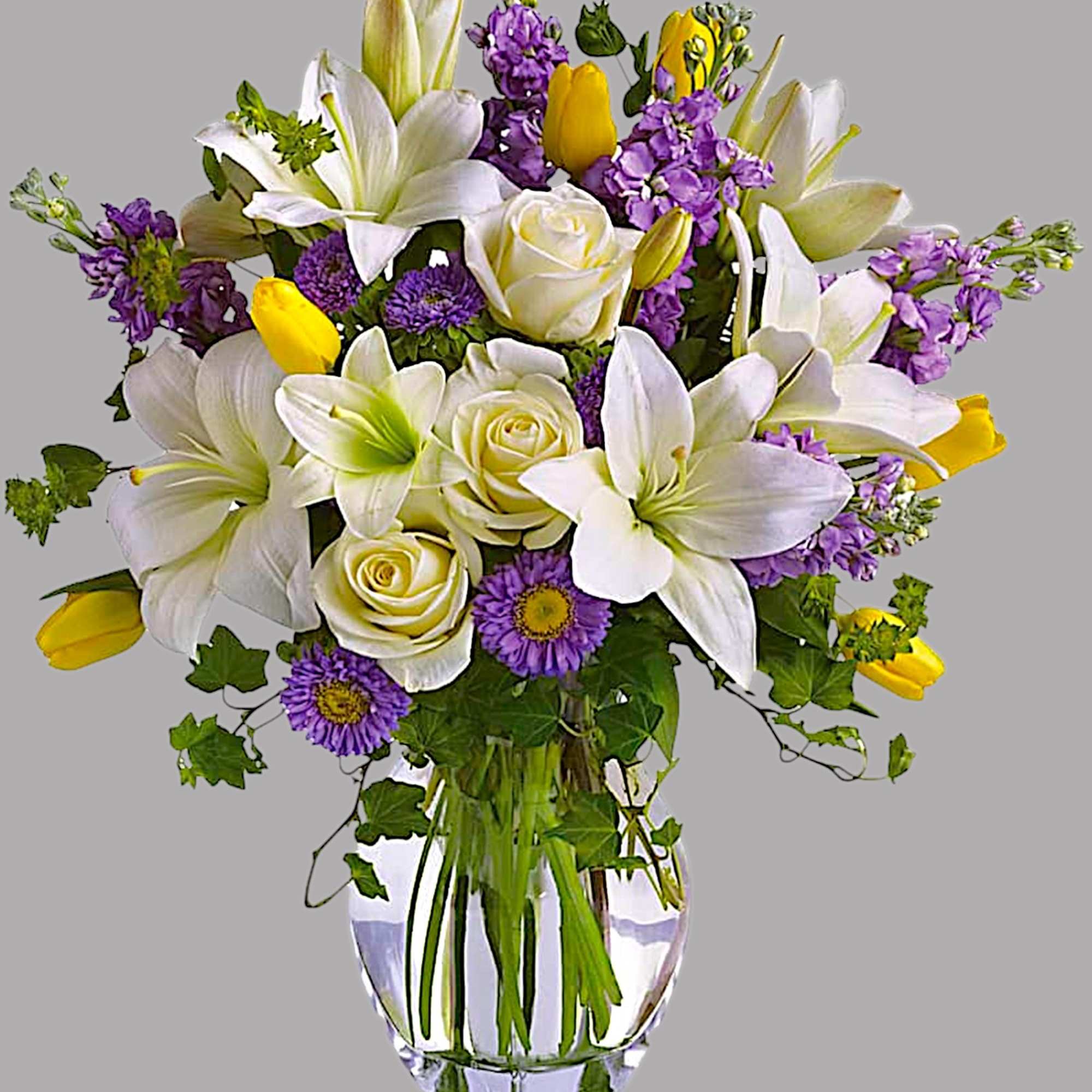 Bouquet of white lilies, cream roses, purple flowers, and yellow tulips in a glass vase