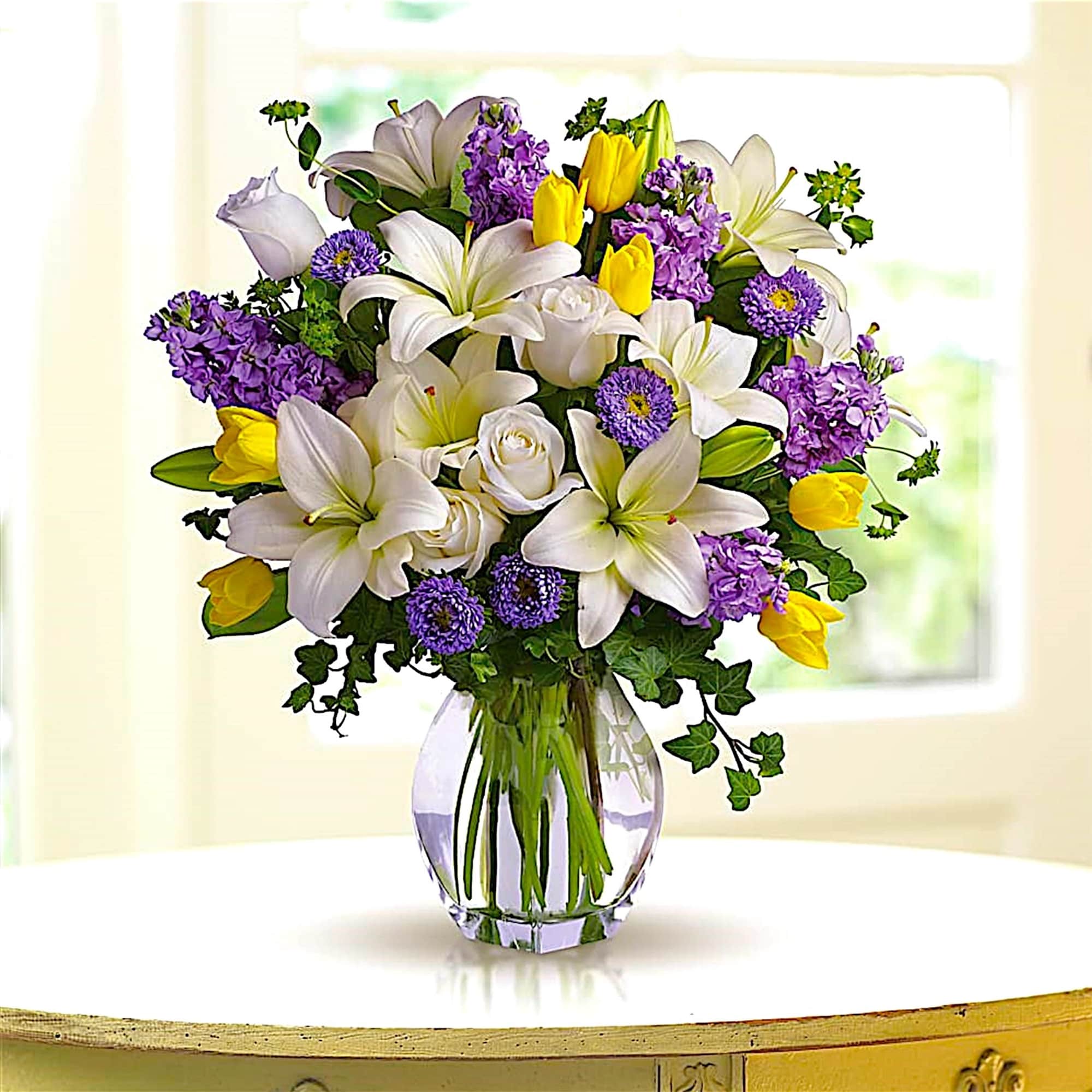 Bouquet of white lilies, yellow tulips, and purple flowers in a glass vase
