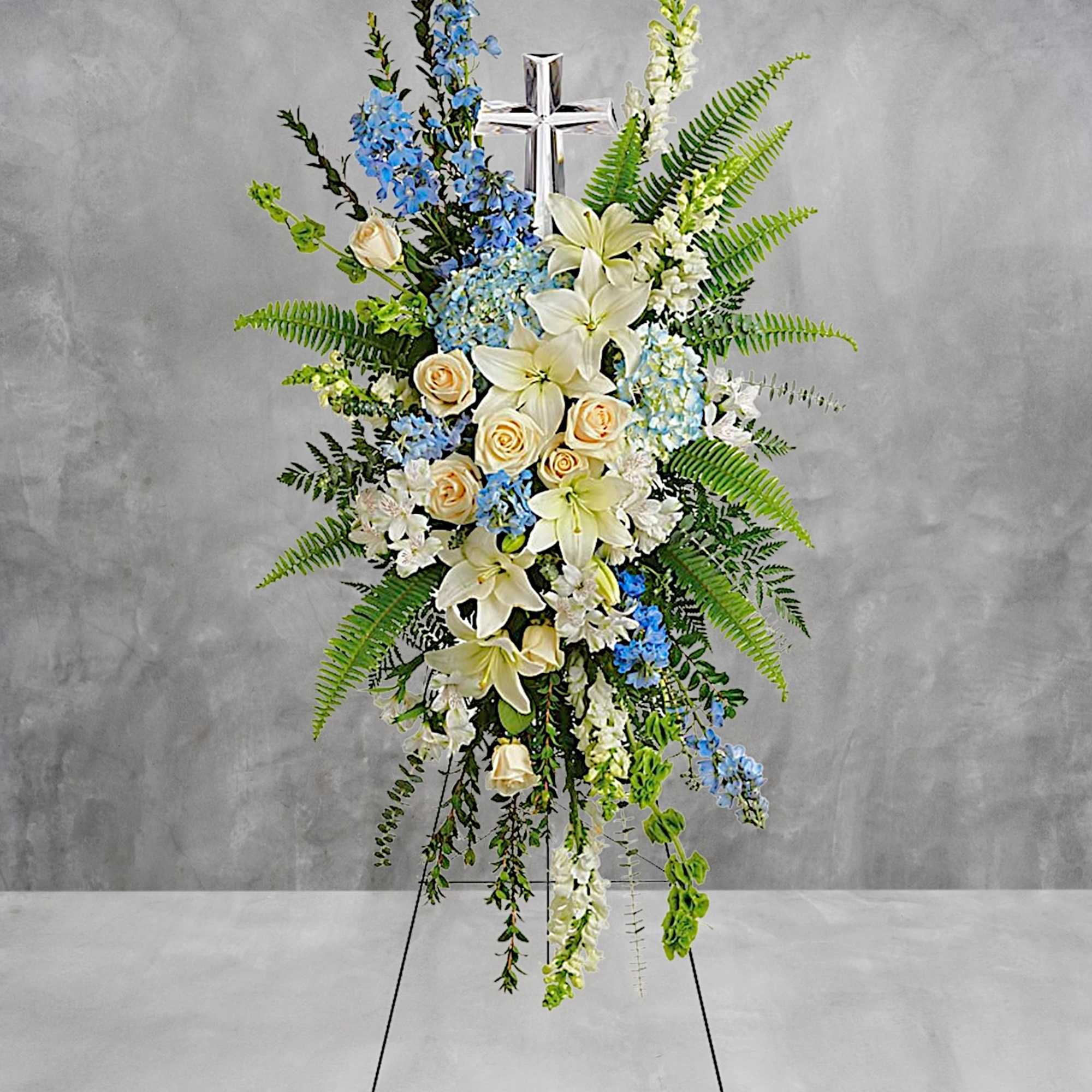 Standing floral spray with white lilies, cream roses, and blue flowers on a cross