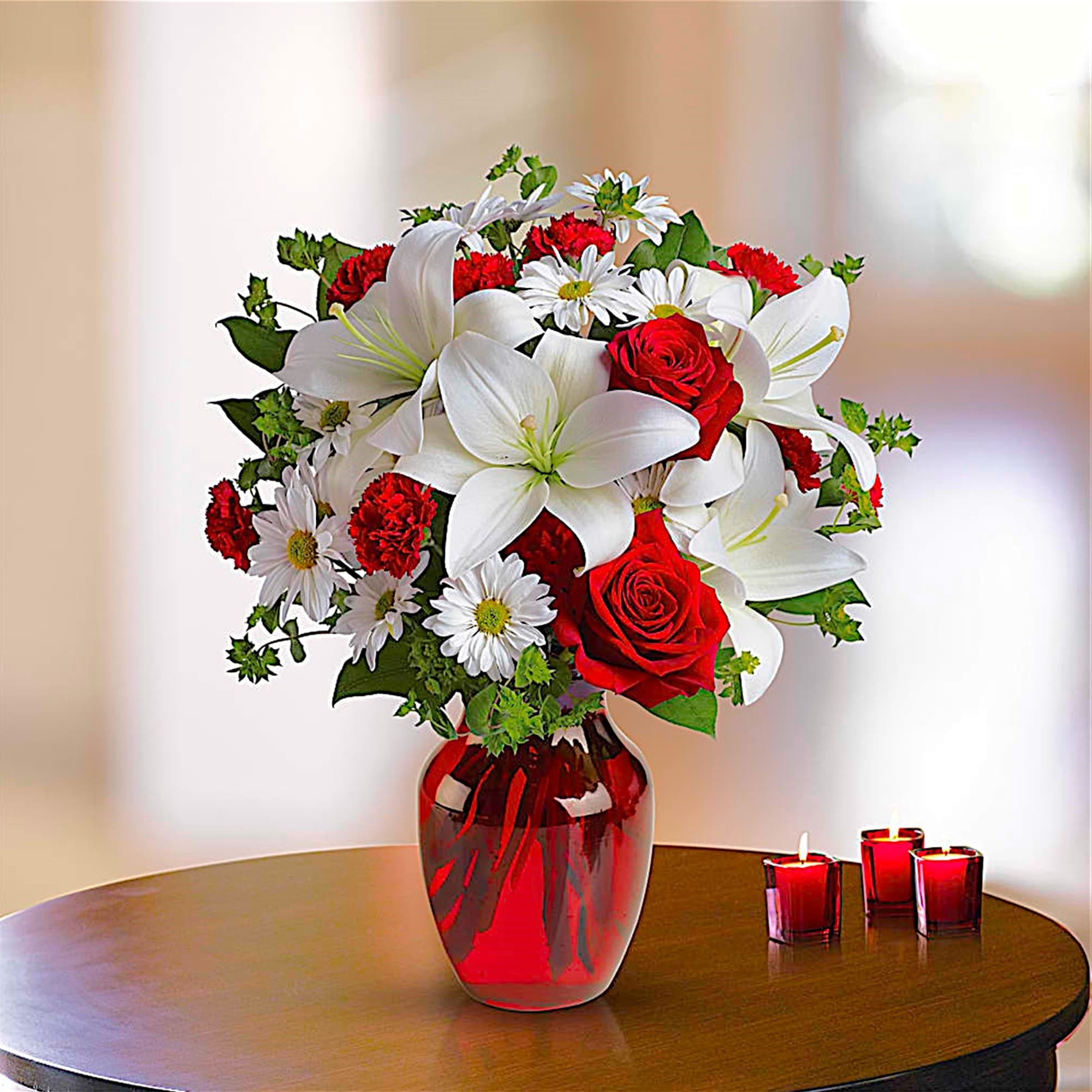 Red and white flower arrangement in a red glass vase with candles nearby