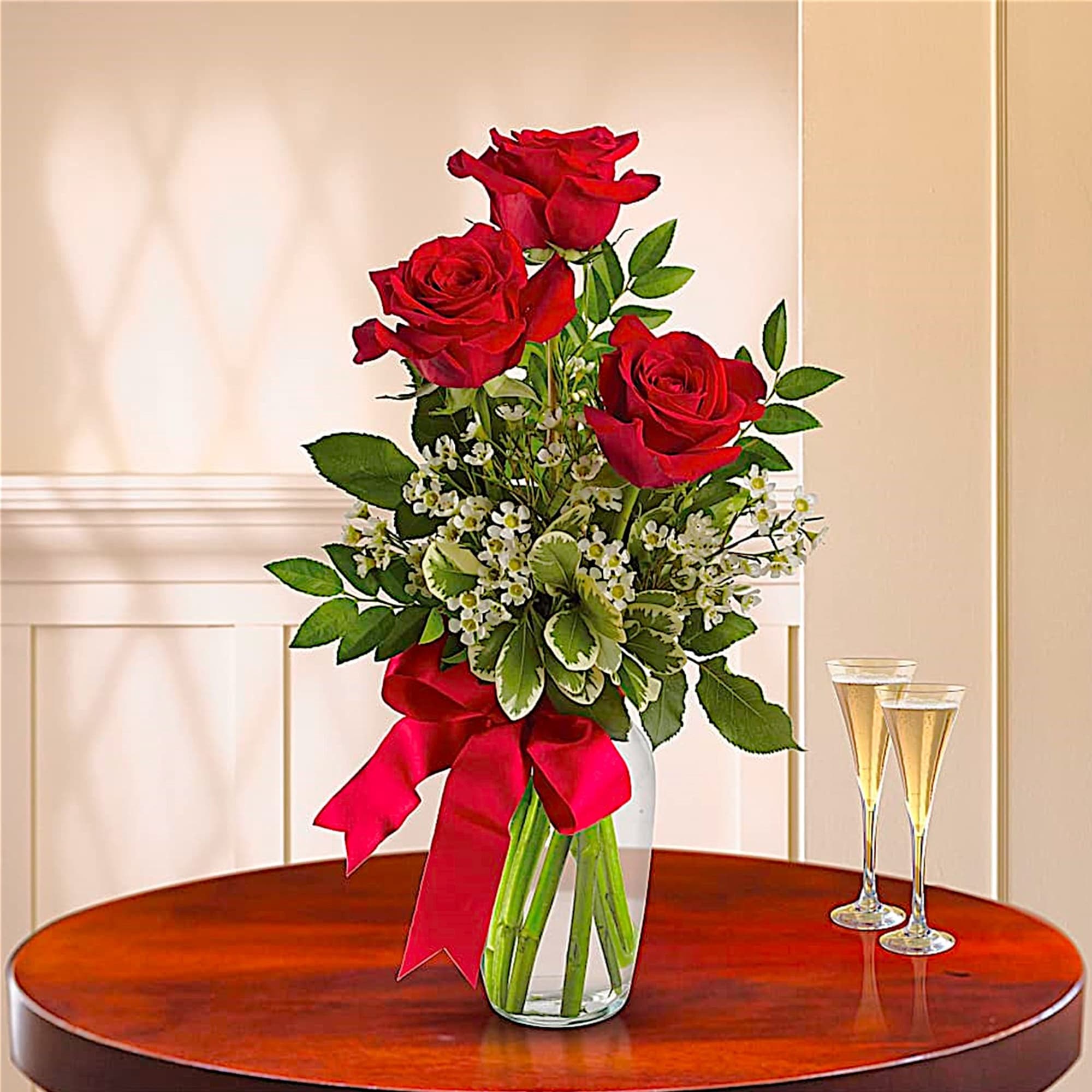 Red roses in a glass vase with a red ribbon, beside two champagne flutes