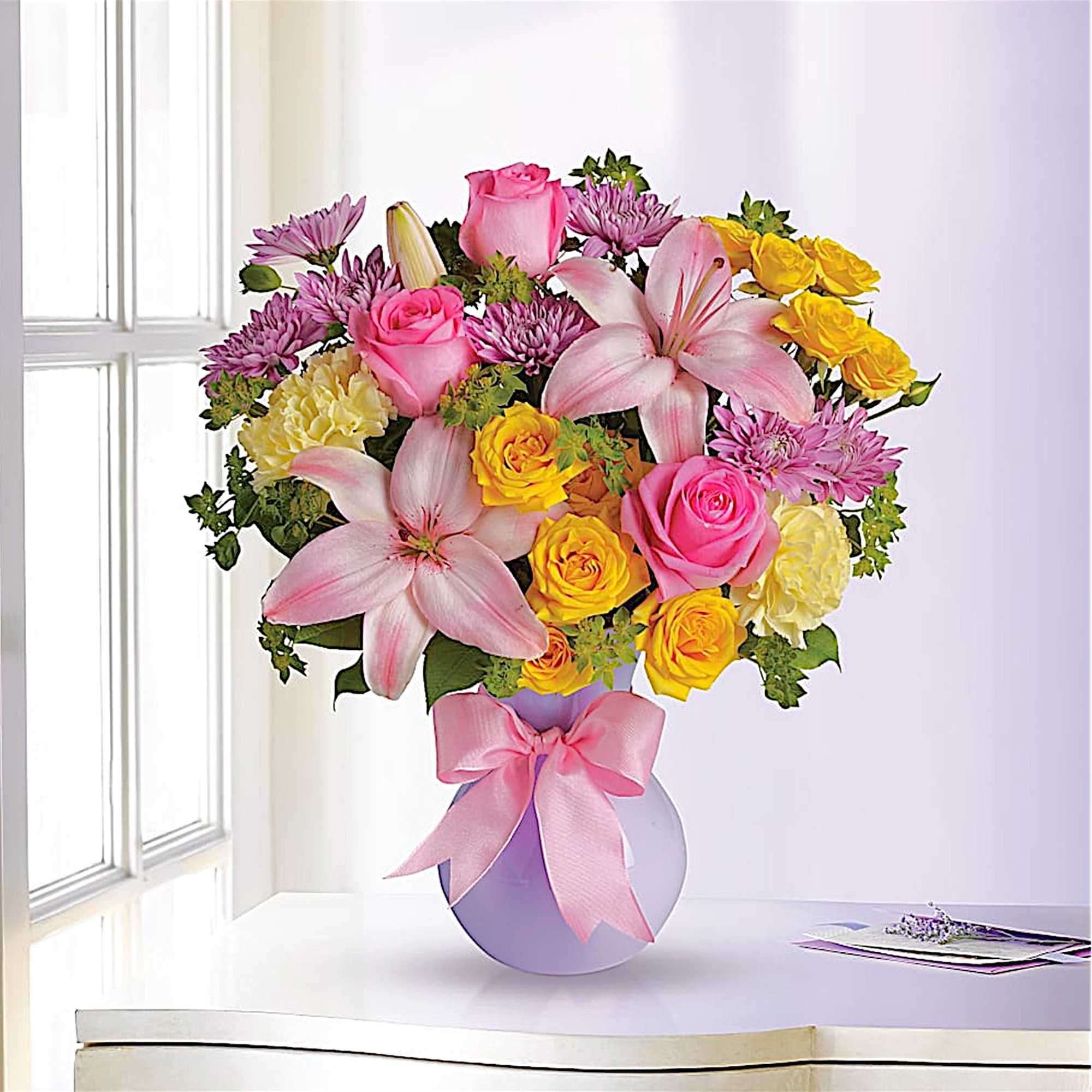 Bouquet of pink lilies, roses, and yellow flowers in a round vase with a pink ribbon