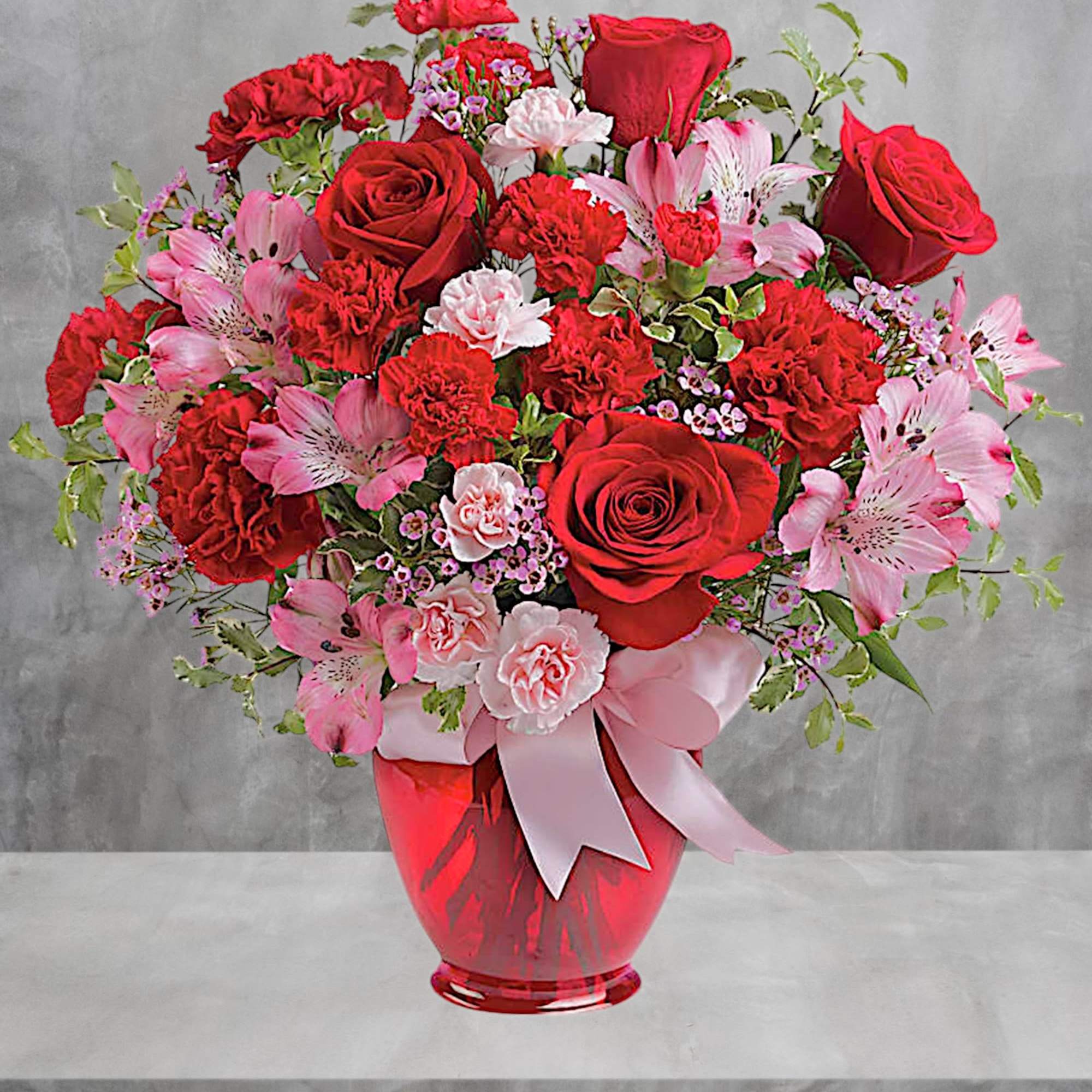 Red roses and pink flowers arranged in a red vase with a pink ribbon