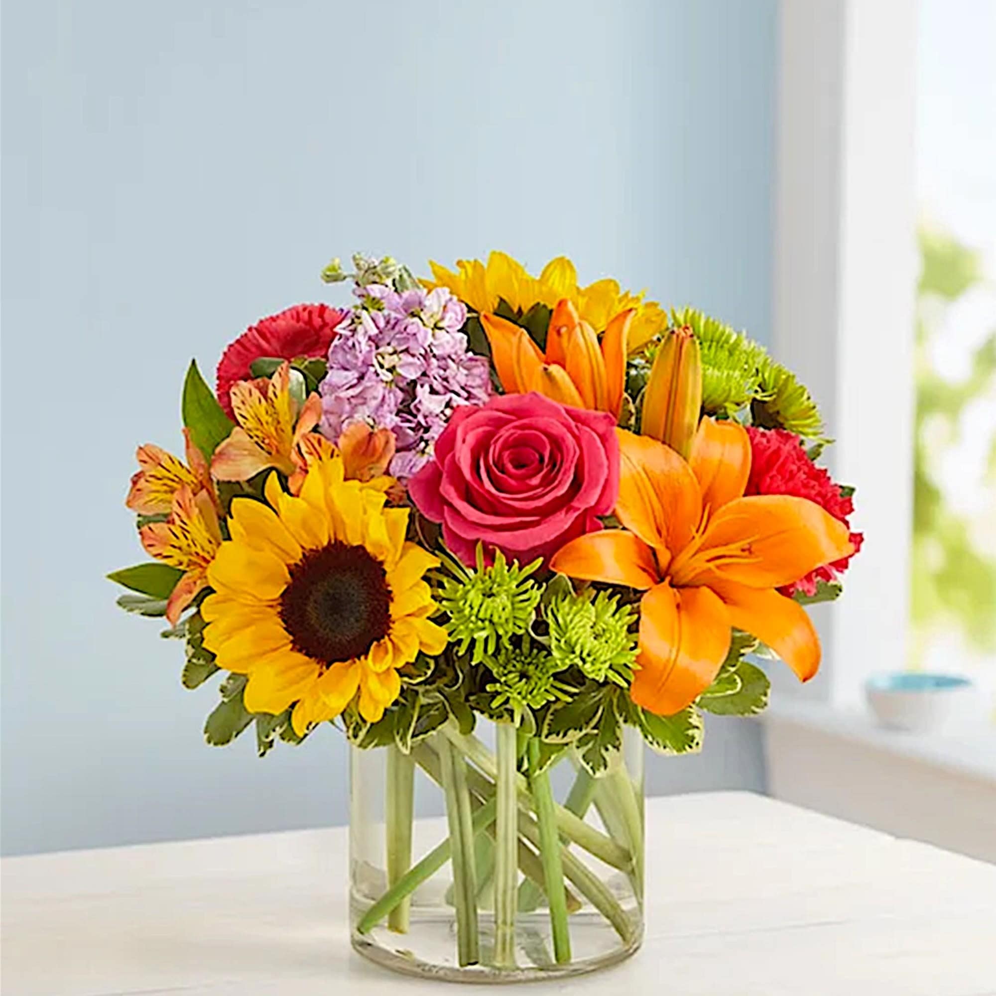 Mixed bouquet with sunflower, rose, lily, and orange blooms in a glass vase