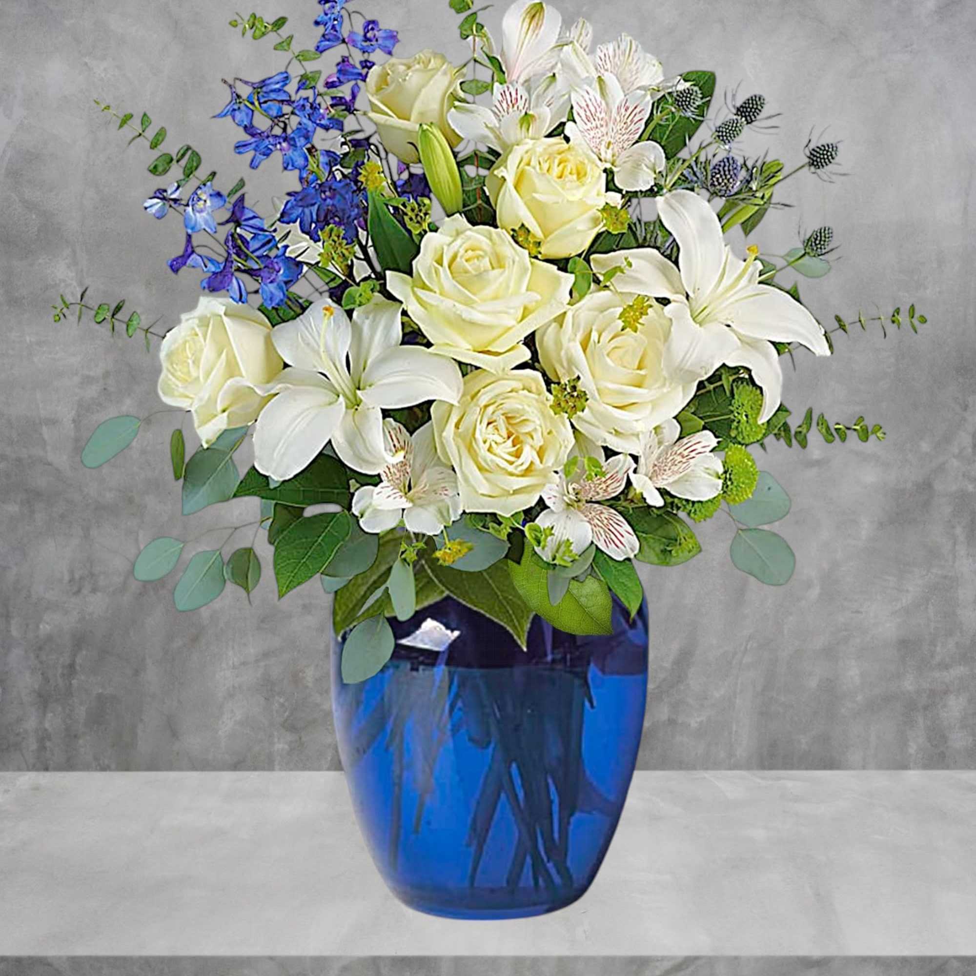 White roses and lilies arranged in a blue glass vase with blue flowers