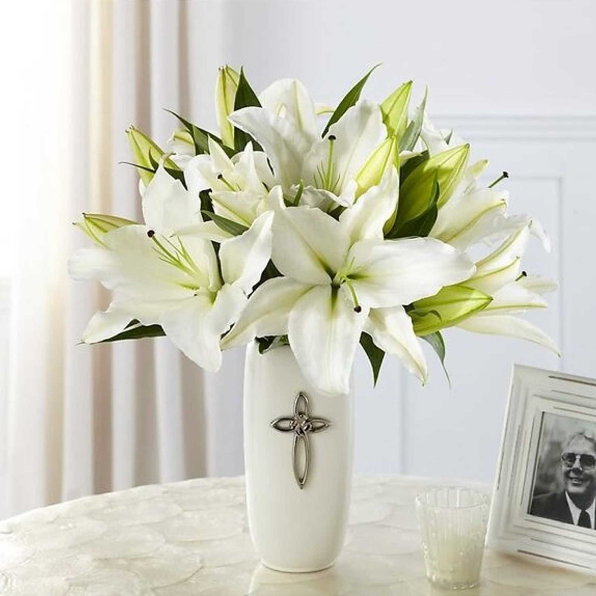 White lilies arranged in a tall white vase with a cross emblem