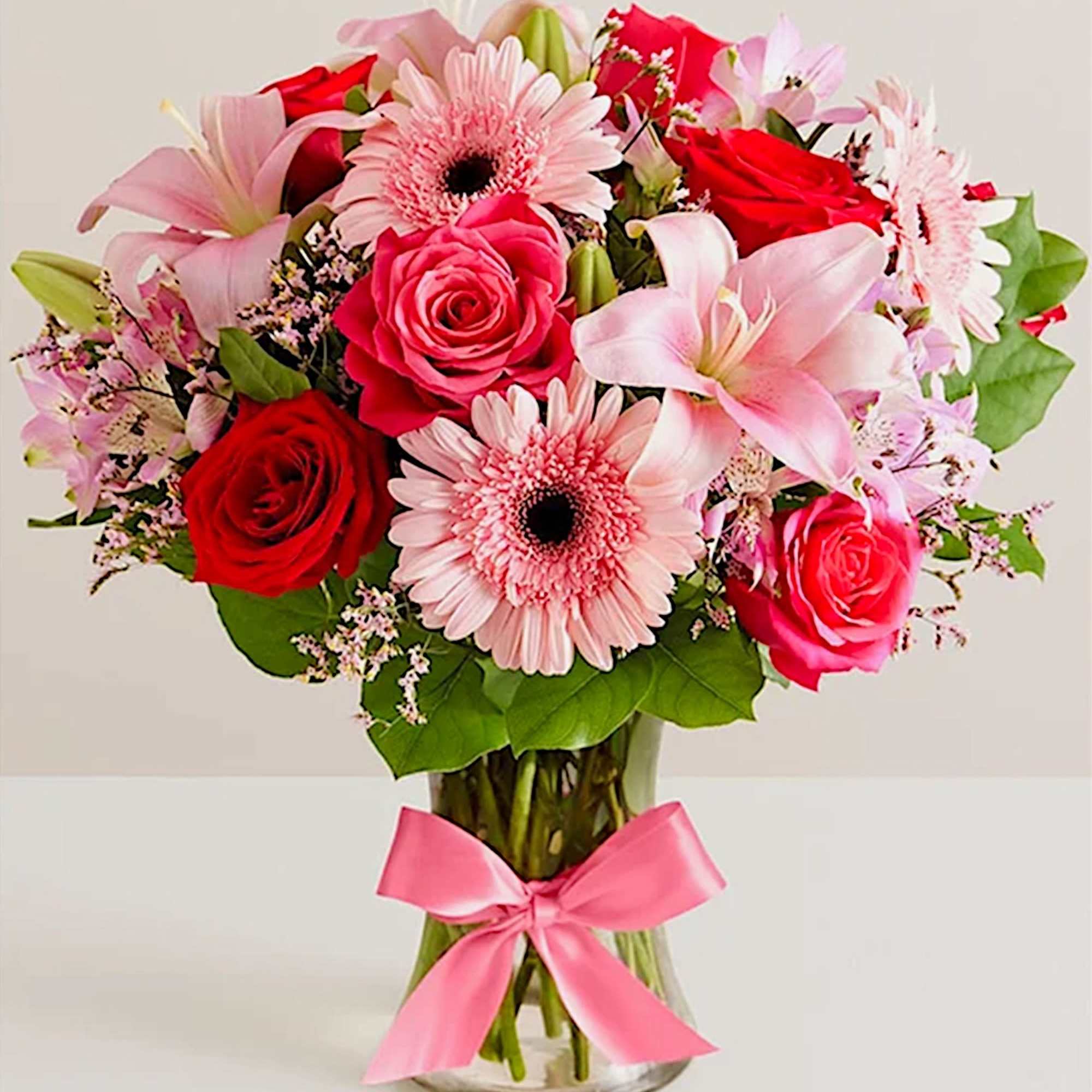 Bouquet of pink lilies, roses, and gerbera daisies in a glass vase with a pink ribbon