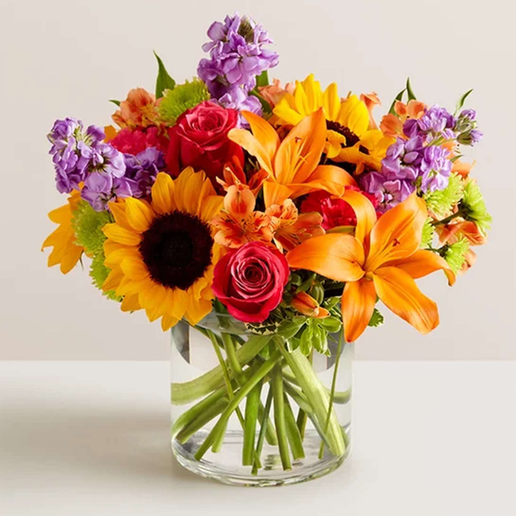 Bright mixed bouquet with sunflowers, roses, lilies, and purple blooms in a glass vase