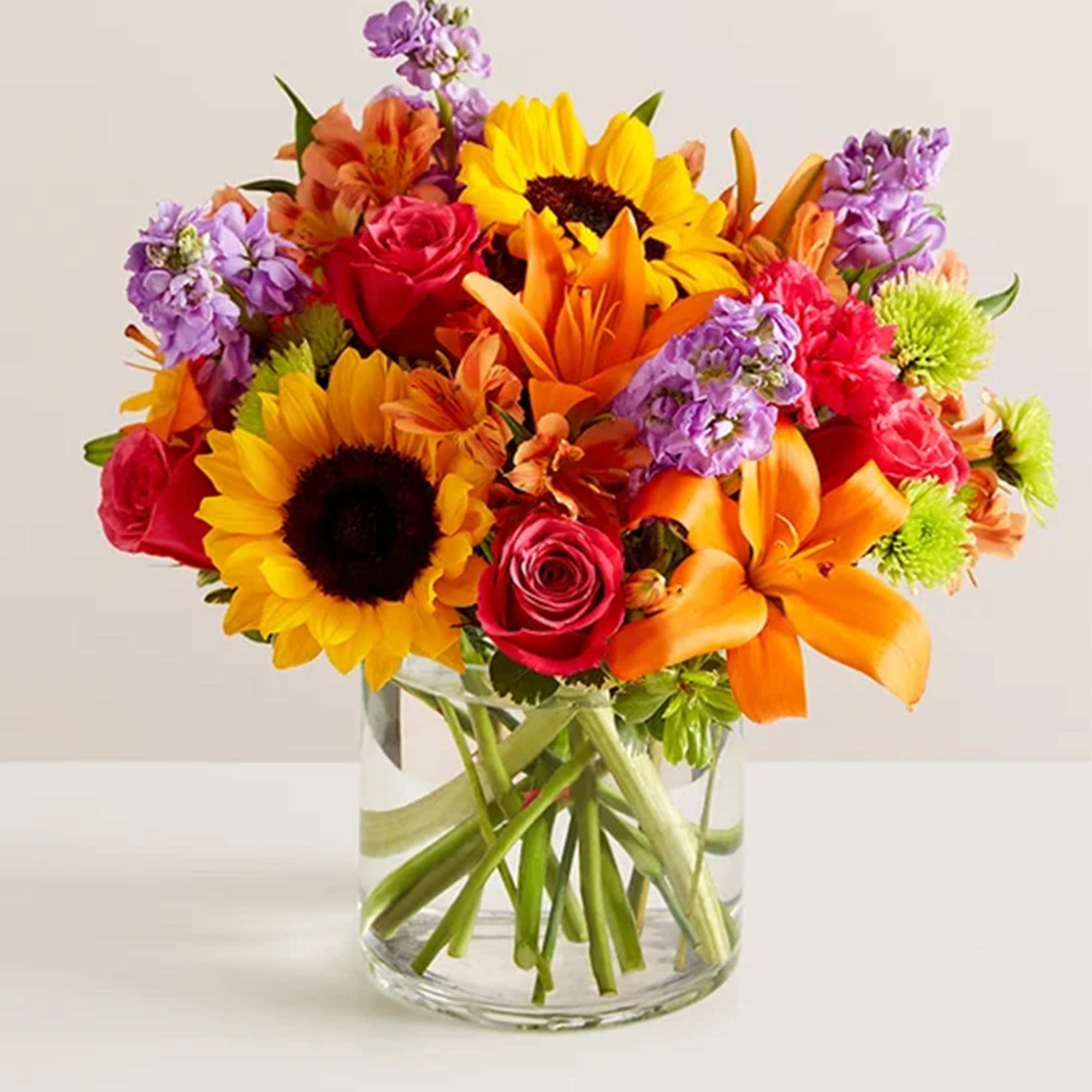 Bright mixed bouquet with sunflowers, roses, lilies, and purple blooms in a glass vase