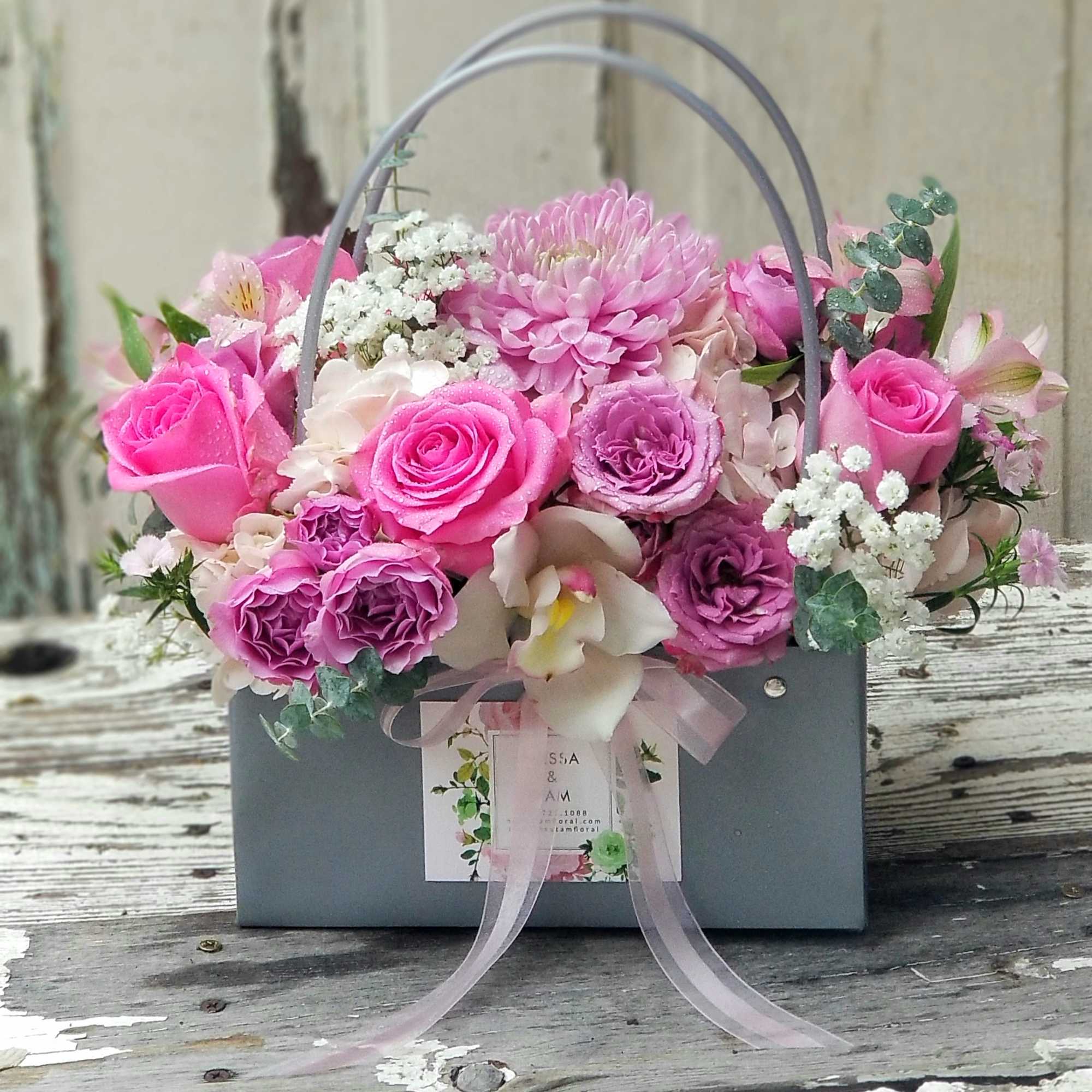 Pink and lavender arrangement with roses, chrysanthemum, and orchid in a gray handled box.