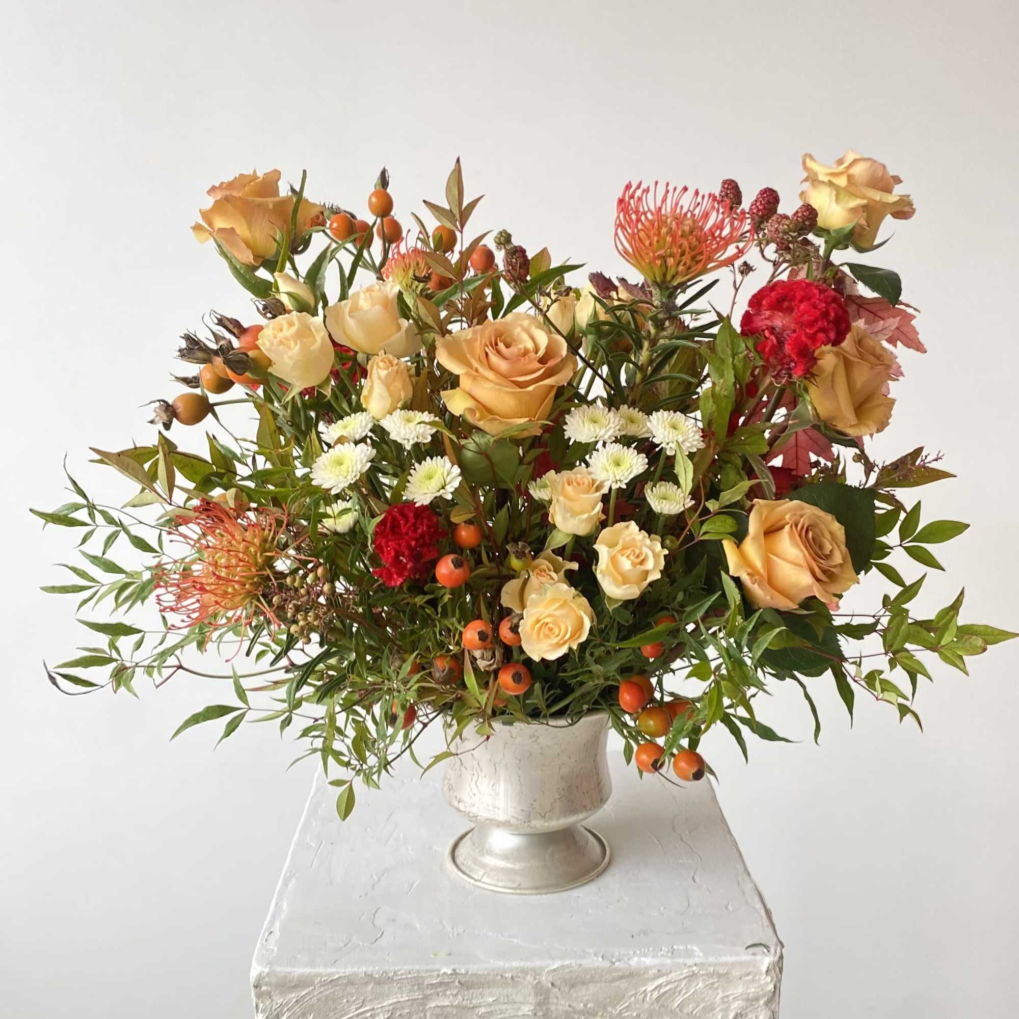 Mixed bouquet of peach roses, white mums, and orange flowers in a silver vase