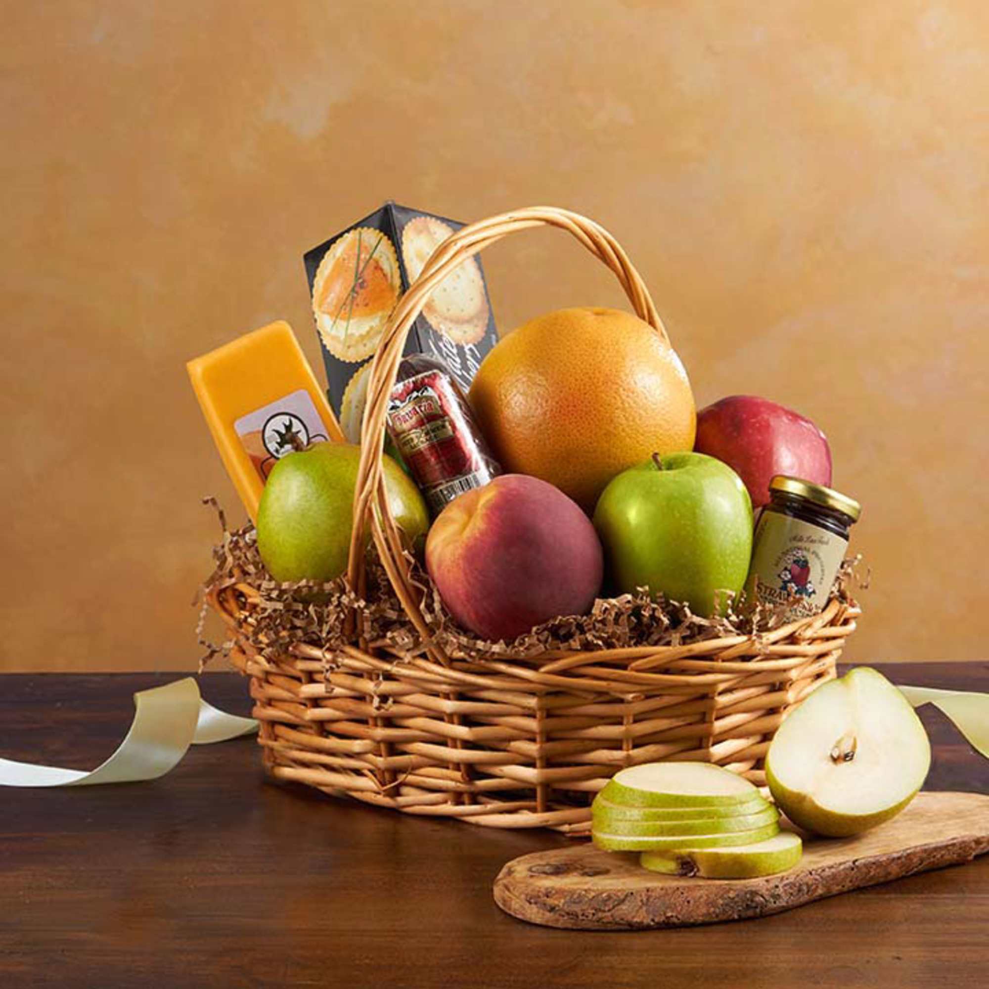 Wicker gift basket with assorted fruit, cheese, sausage, crackers, and jam beside a sliced pear.