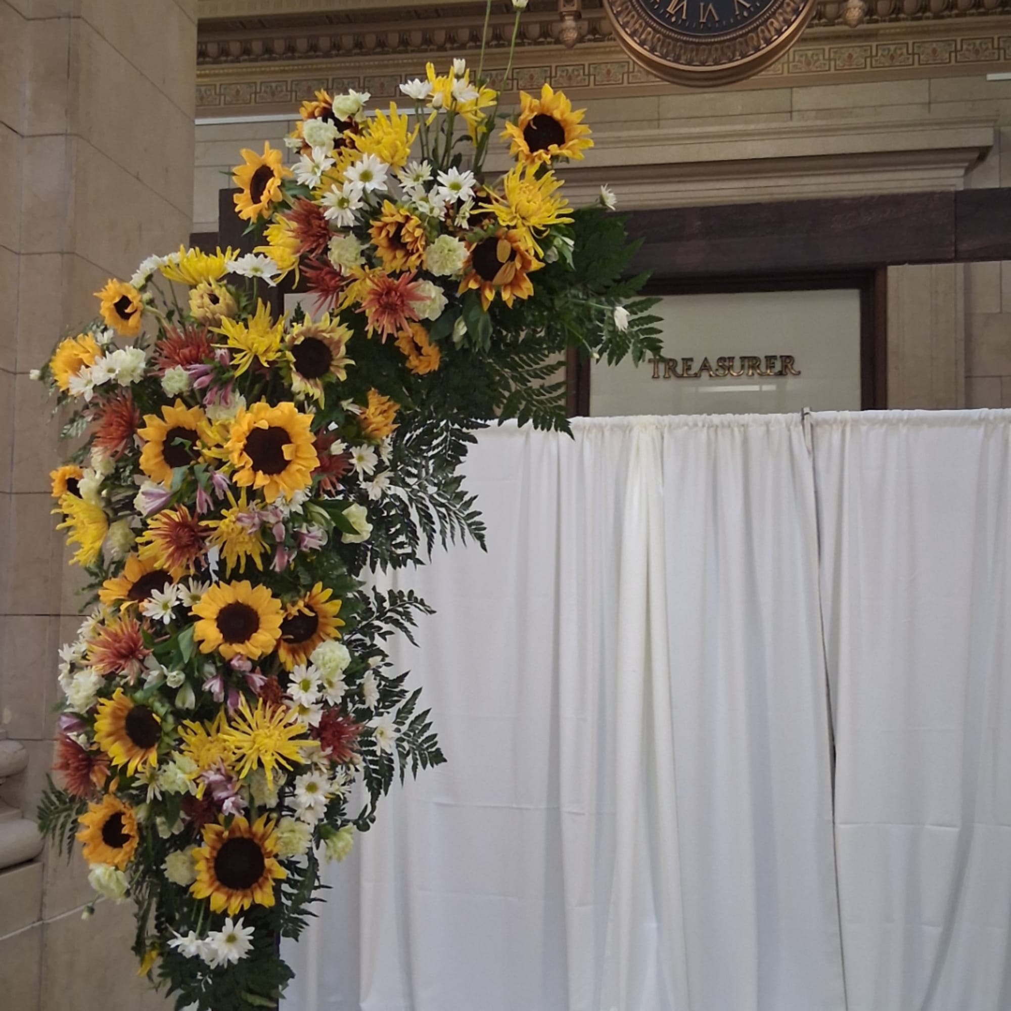 Large floral arch with sunflowers and mixed blooms beside white draping