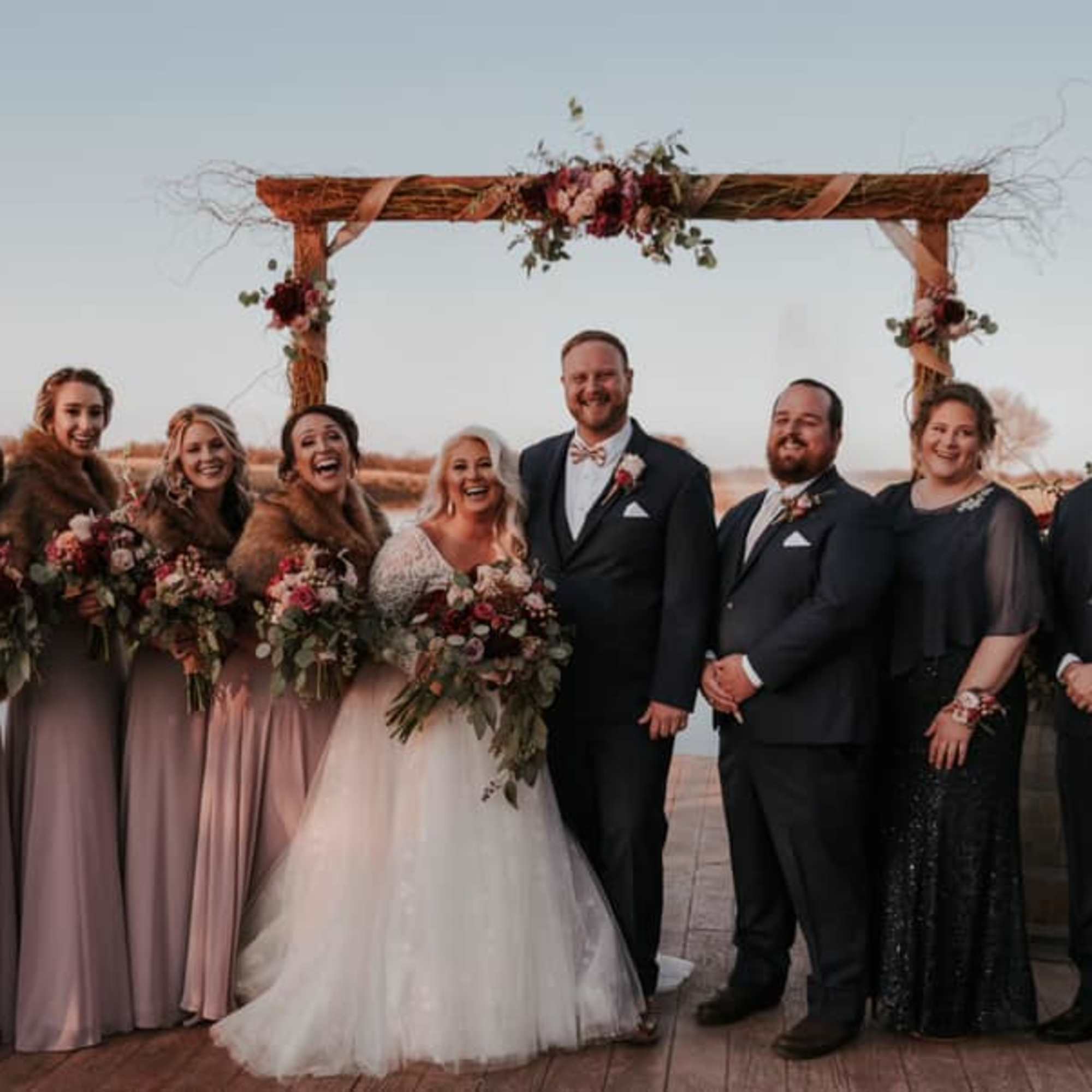 Wedding party posing with bouquets under a floral arch by the water