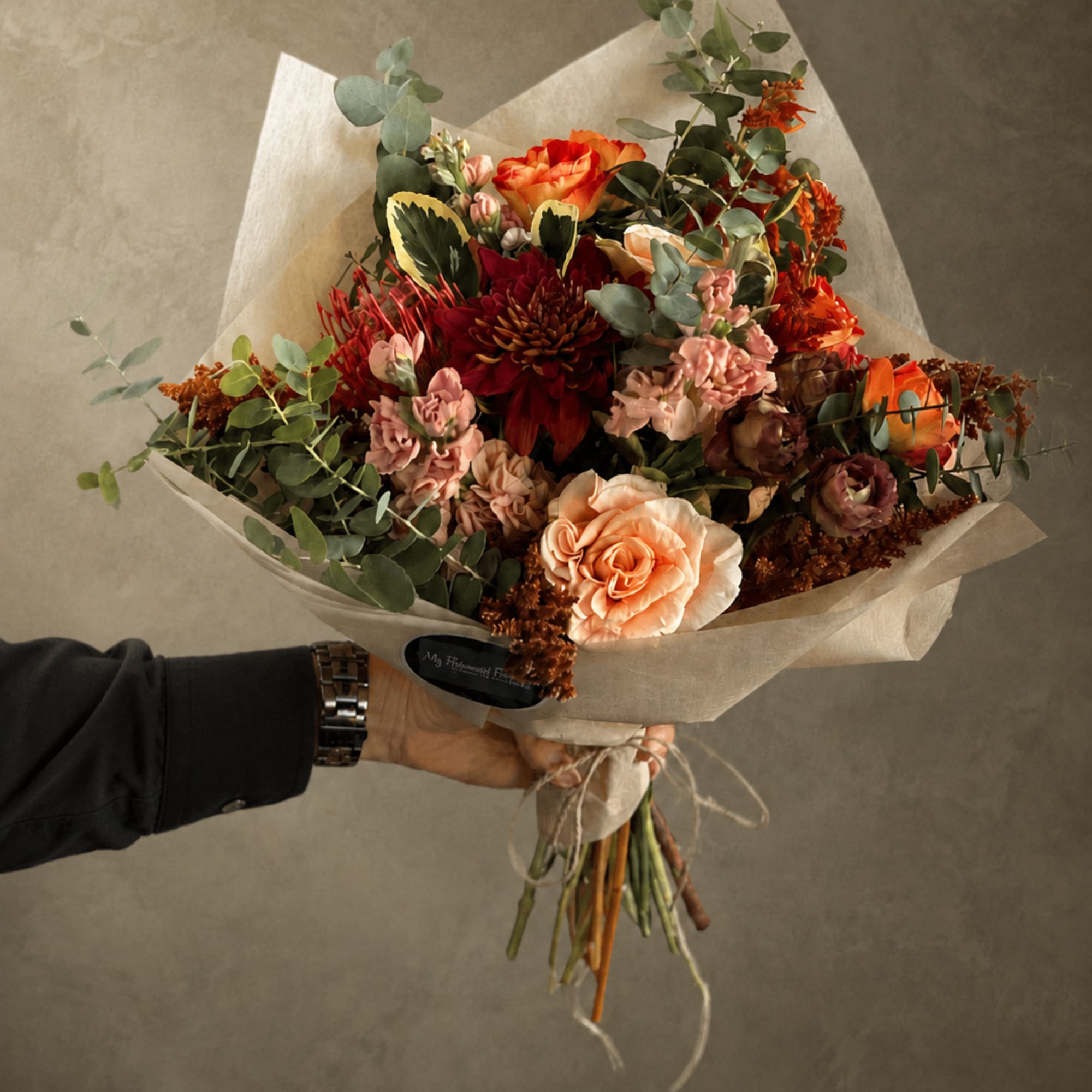 Handheld bouquet of orange, peach, and burgundy flowers wrapped in paper