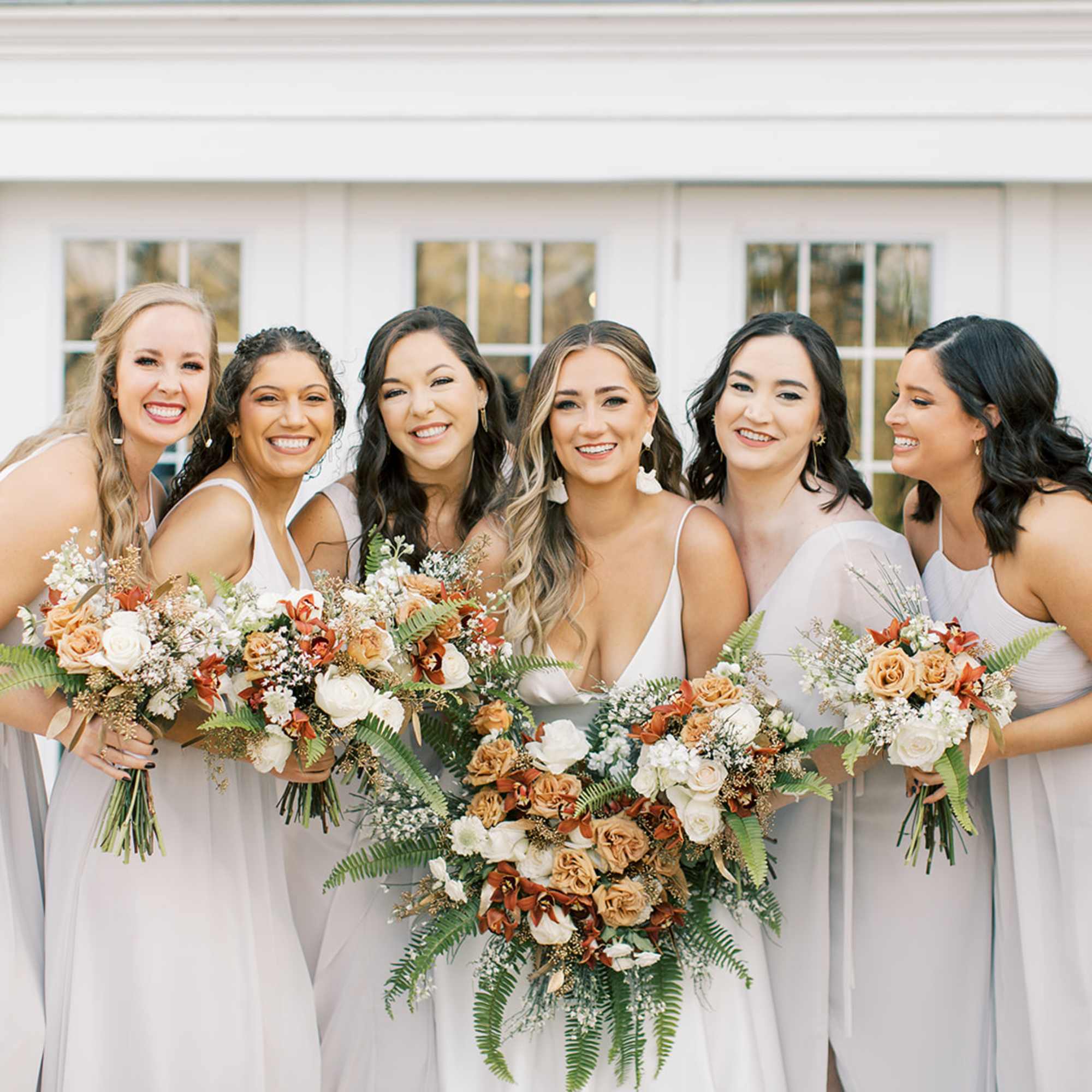 Six women in light dresses holding peach and white bouquets.