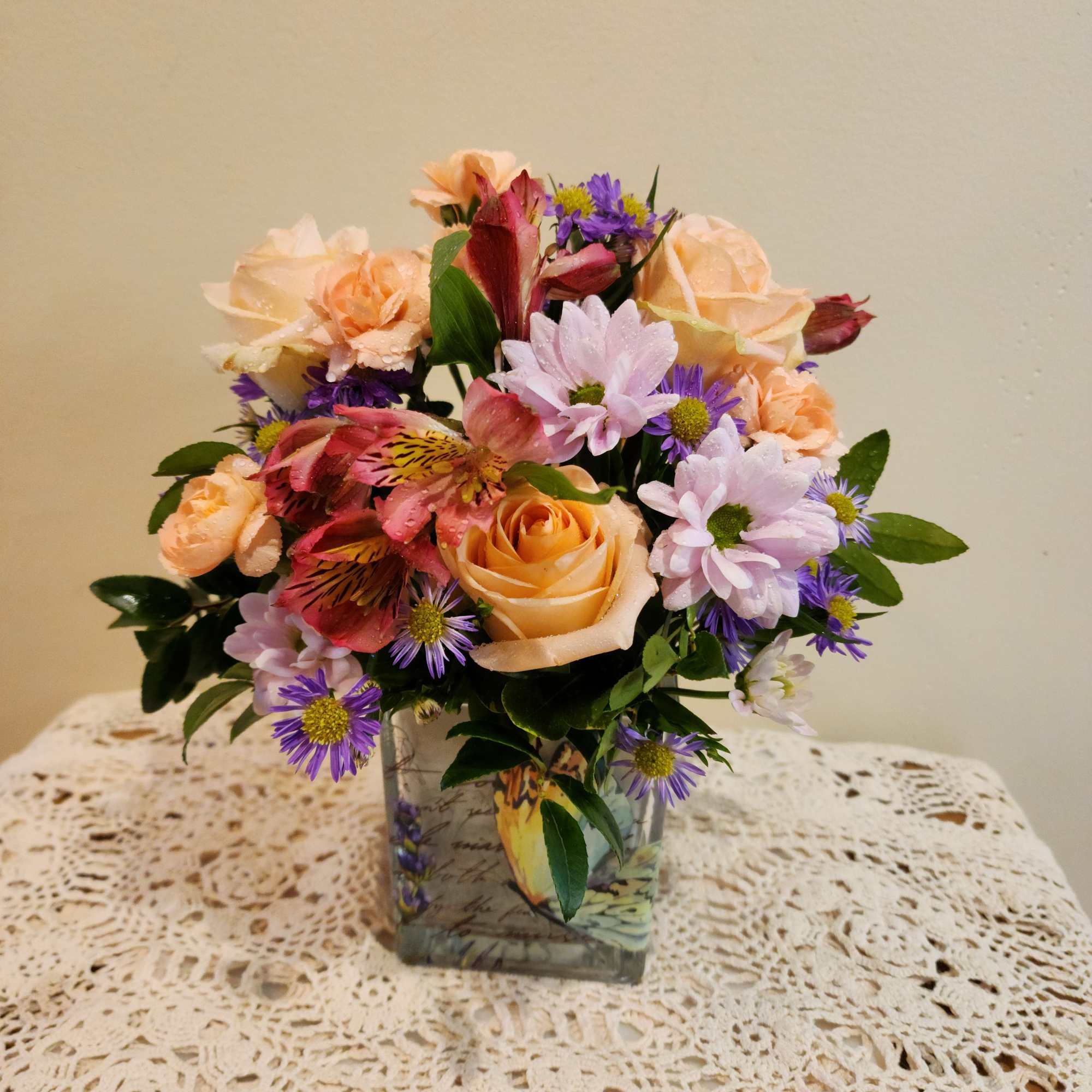 Mixed bouquet of peach roses, pink daisies, and purple asters in a square glass vase