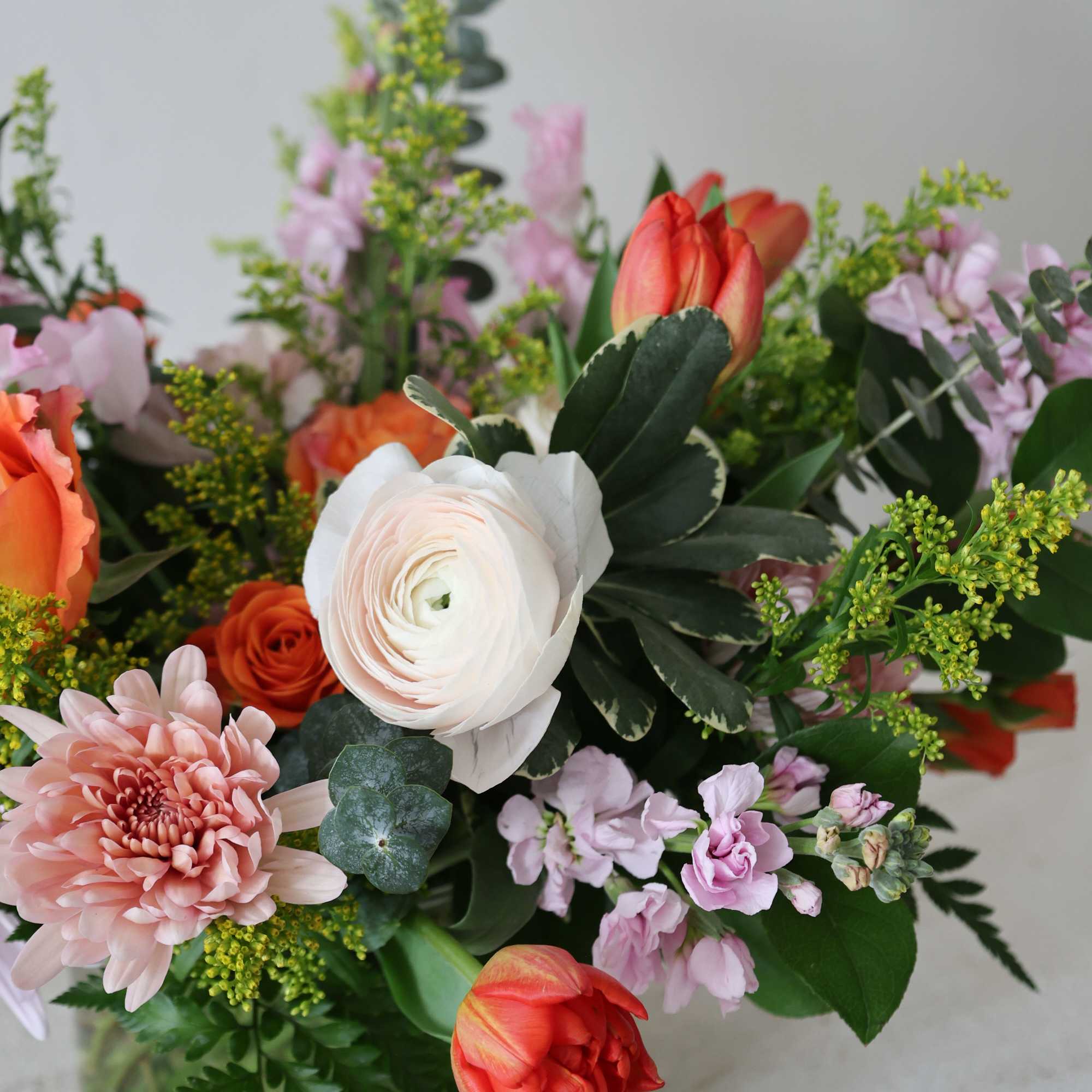 Mixed bouquet with orange tulips, white ranunculus, and pink blooms in a glass vase
