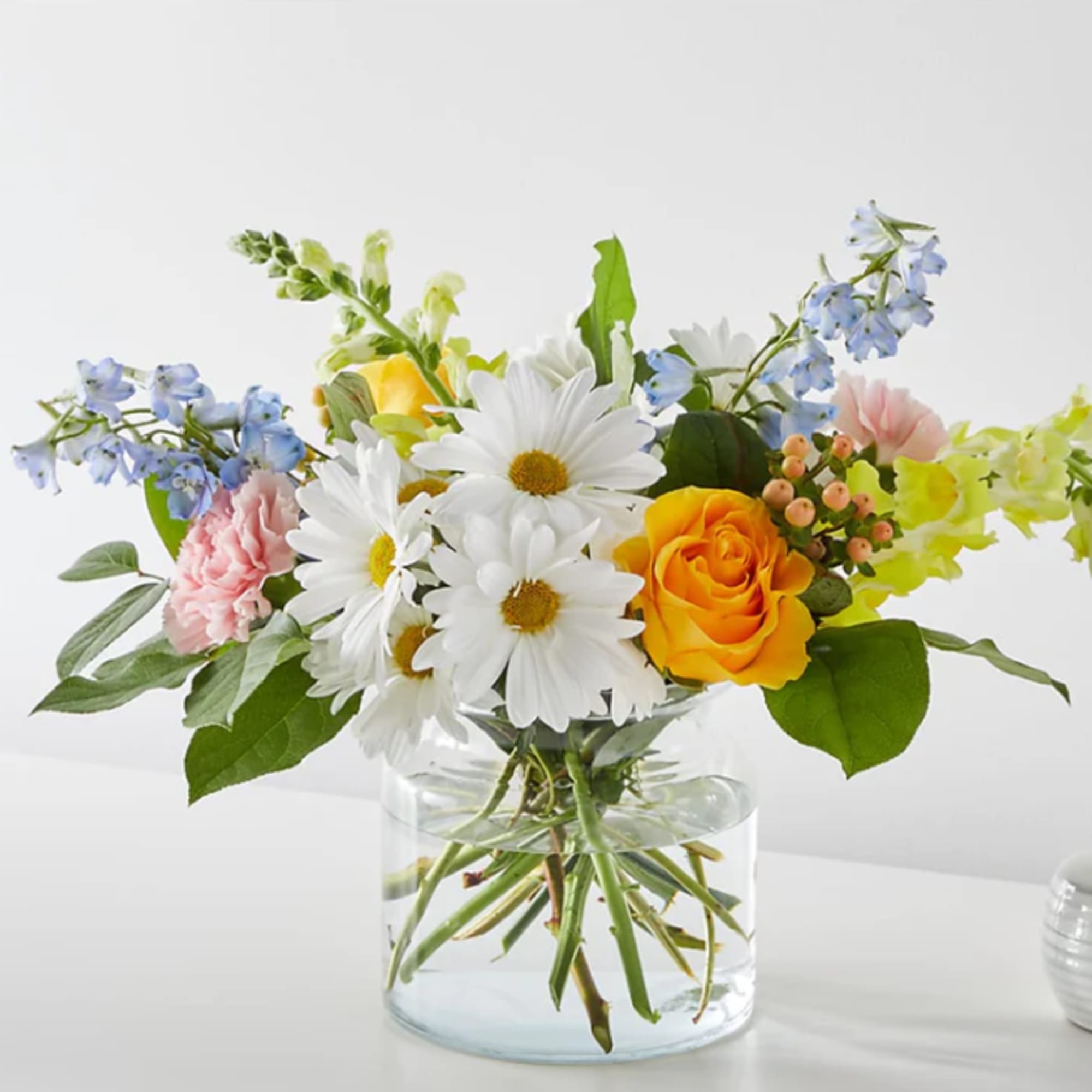 Bouquet of white daisies, yellow roses, and blue flowers in a clear glass vase