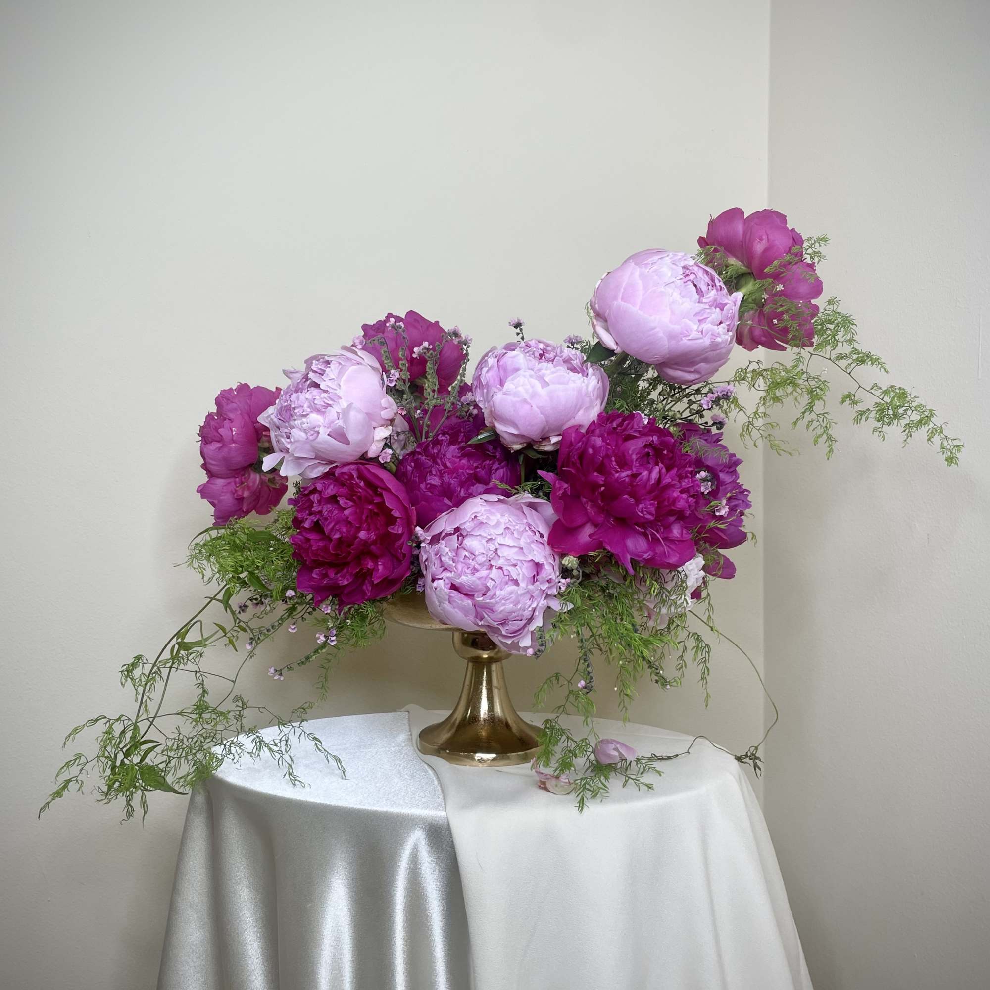 Low compote arrangement of pink and magenta peonies with trailing greenery in a gold pedestal vase.