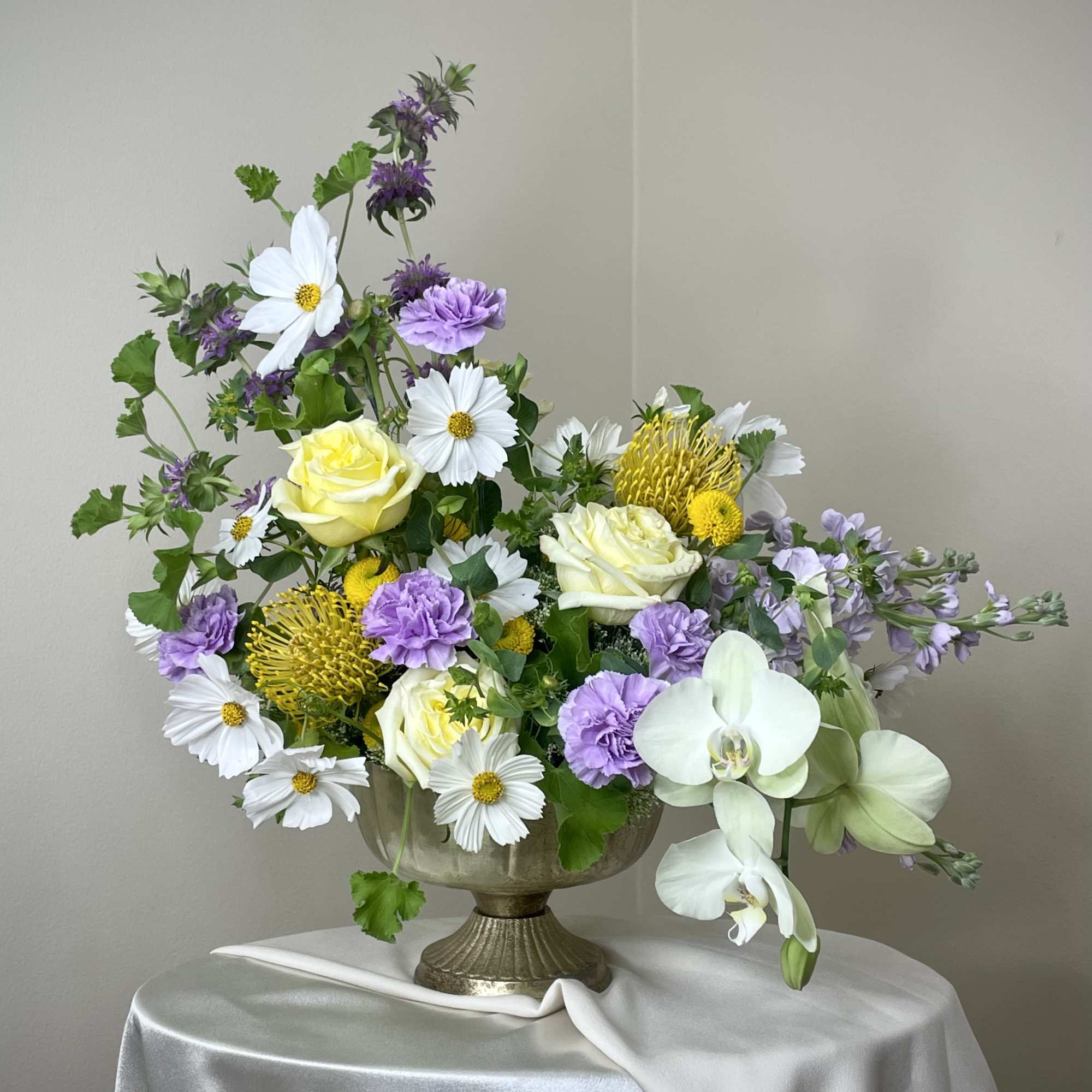 Garden-style arrangement of white, yellow, and lavender flowers in a metal pedestal bowl on a draped table.