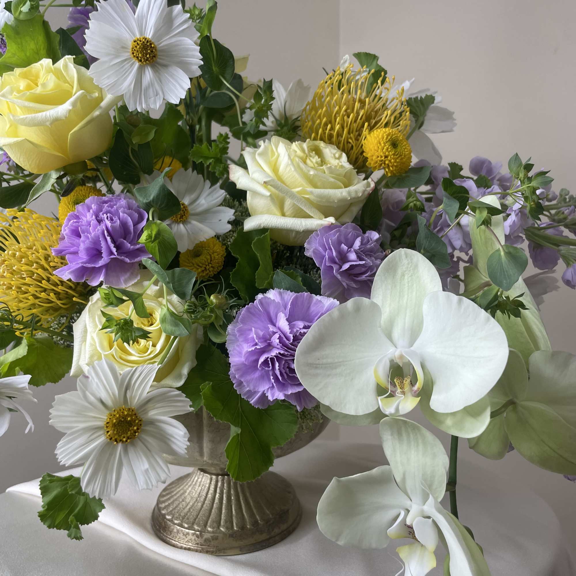 Mixed arrangement of white orchids, yellow roses, and purple flowers in a pedestal vase.