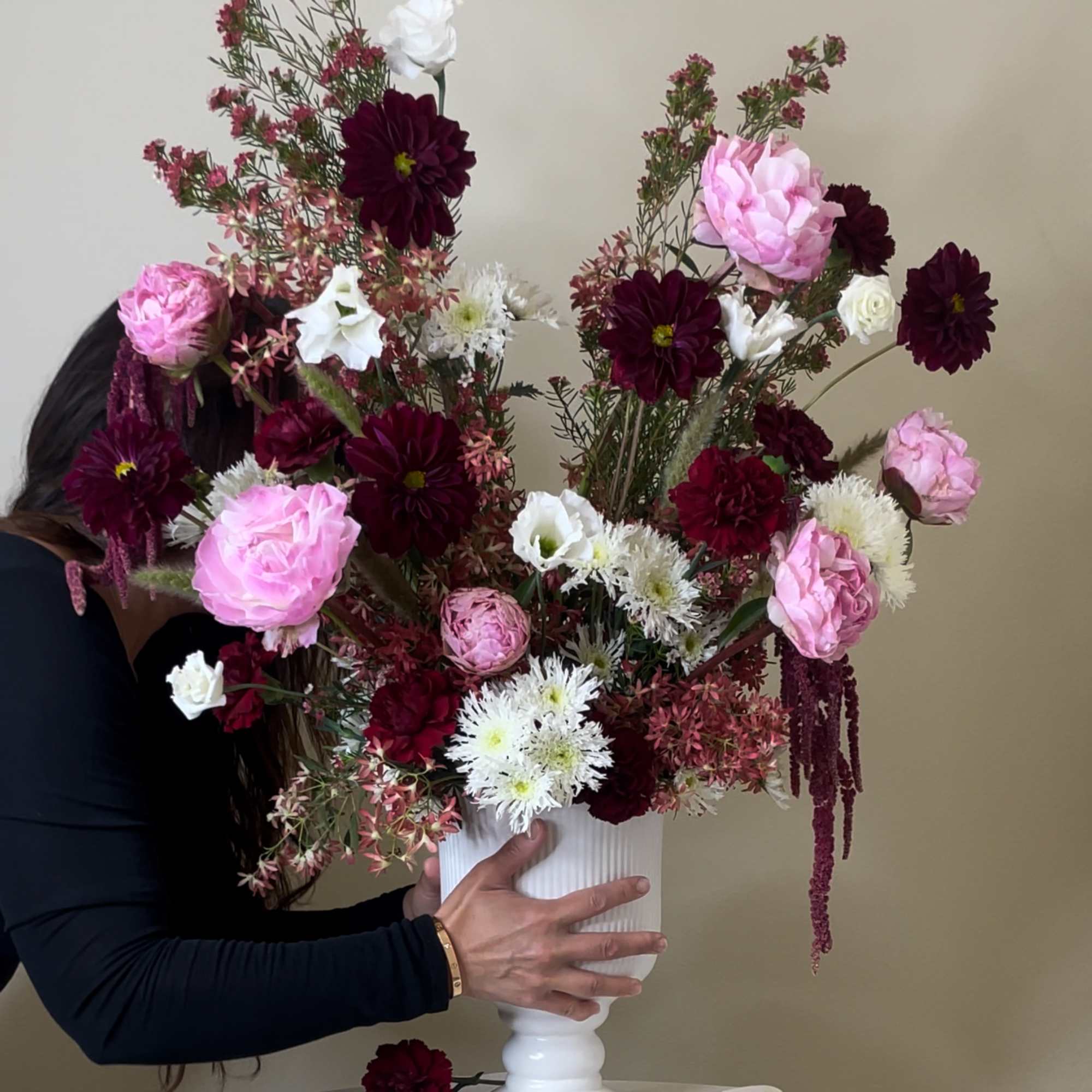 Tall arrangement of burgundy, pink, and white flowers in a white pedestal vase being adjusted by a person.