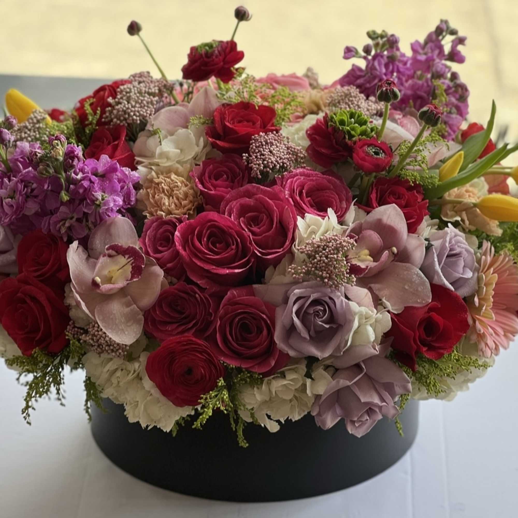 Round arrangement of pink and red roses with purple blooms in a black box