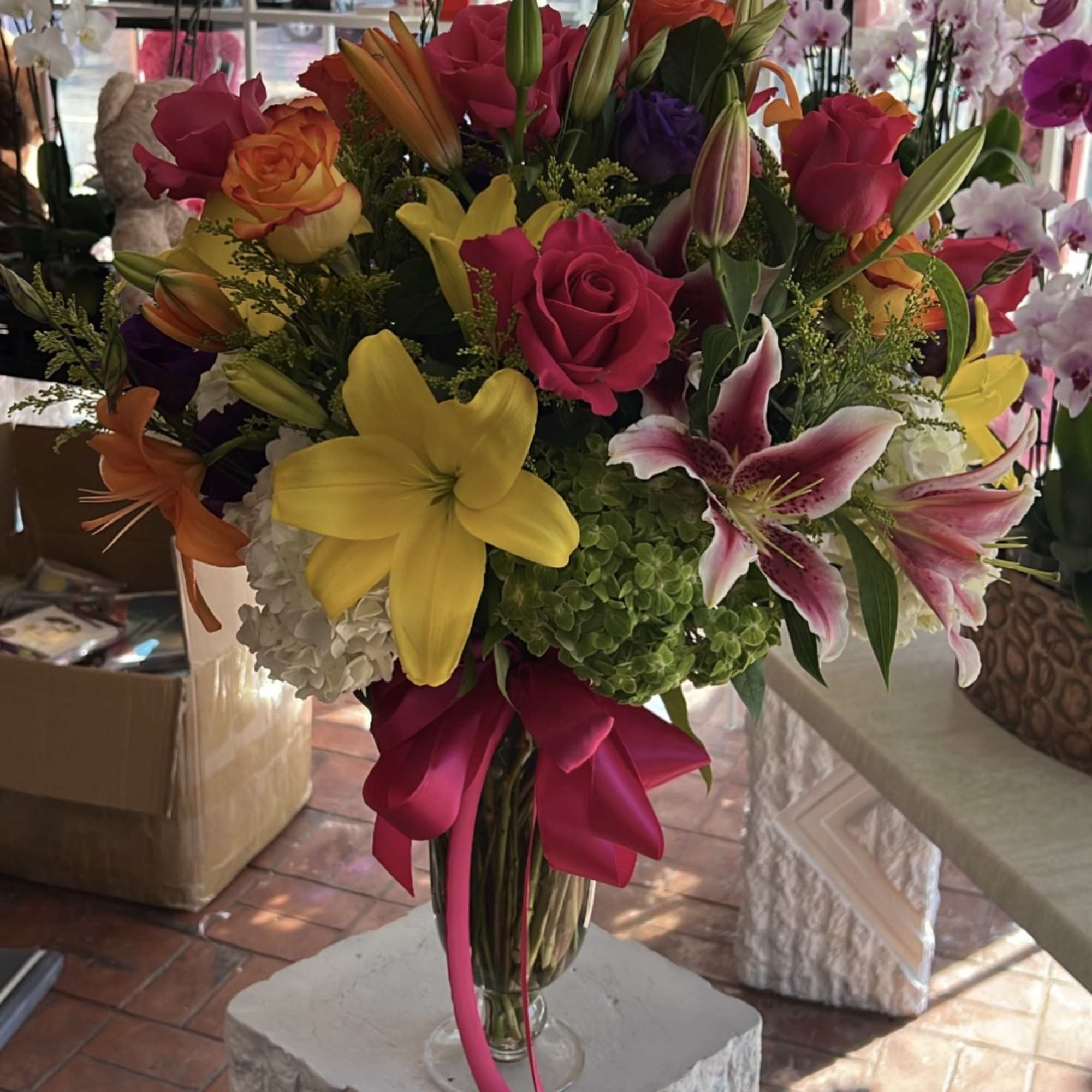 Colorful bouquet of roses, lilies, and orchids in a glass vase with a pink ribbon