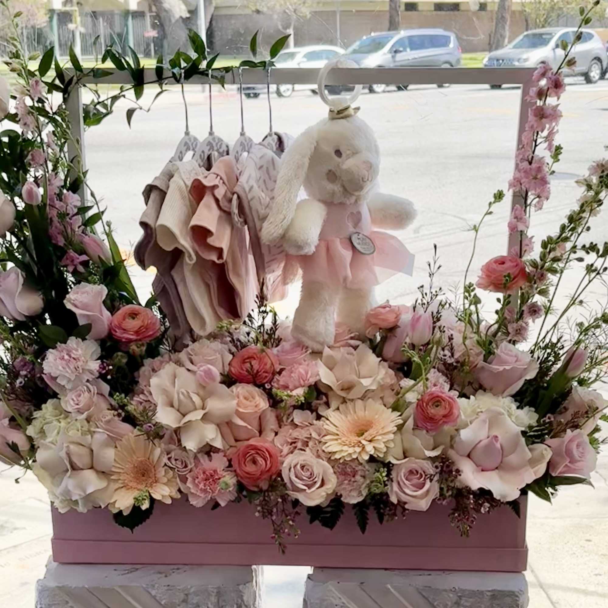 Pink floral display with a teddy bear and baby clothes in a box, photo 2 of 4
