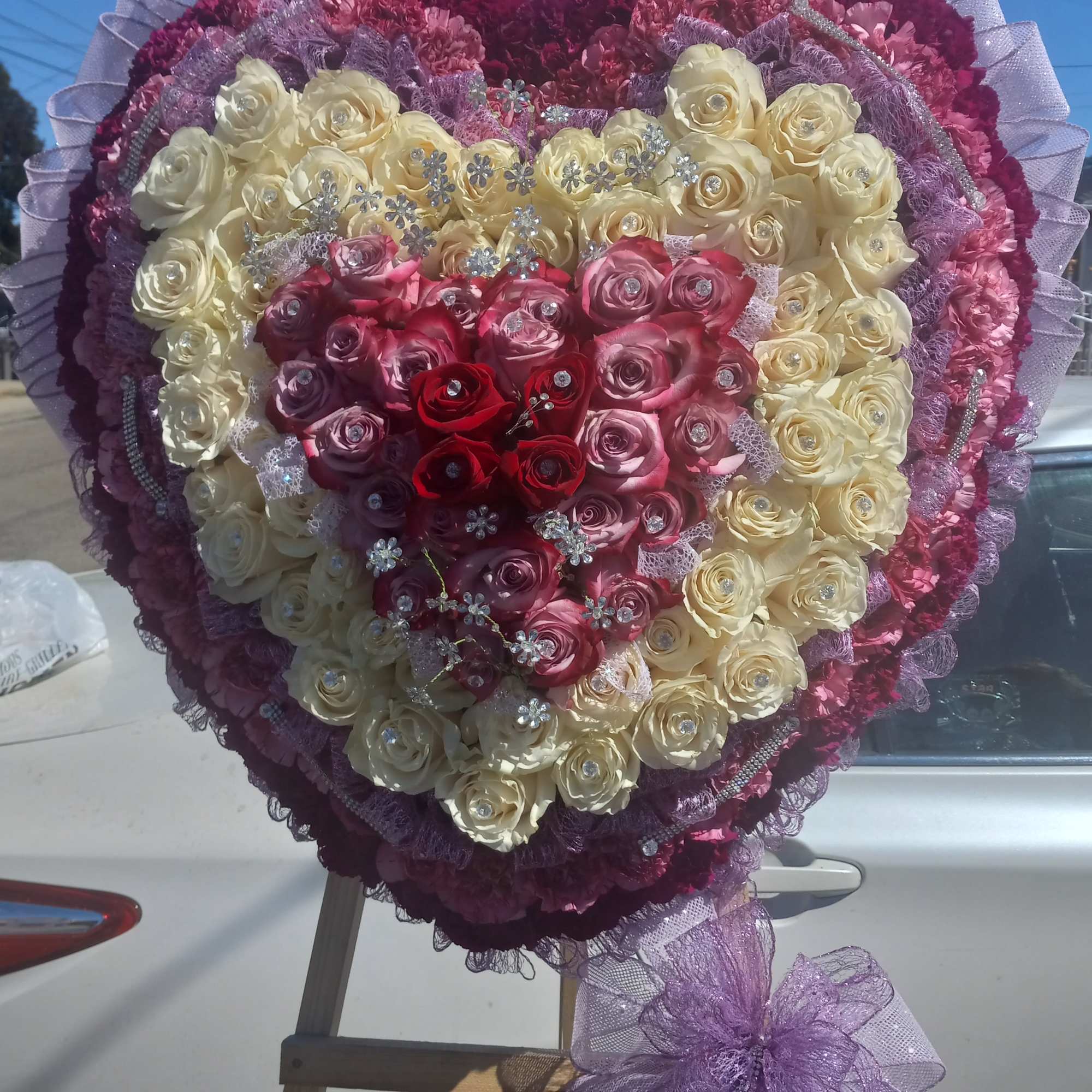 Heart-shaped rose arrangement in cream, pink, and red with purple ribbon trim