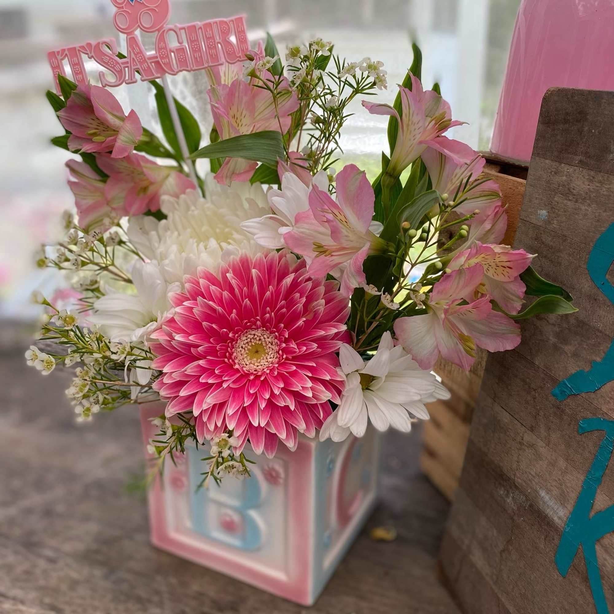 Pink and white baby-themed flower arrangement in a pastel block vase with It's a Girl pick