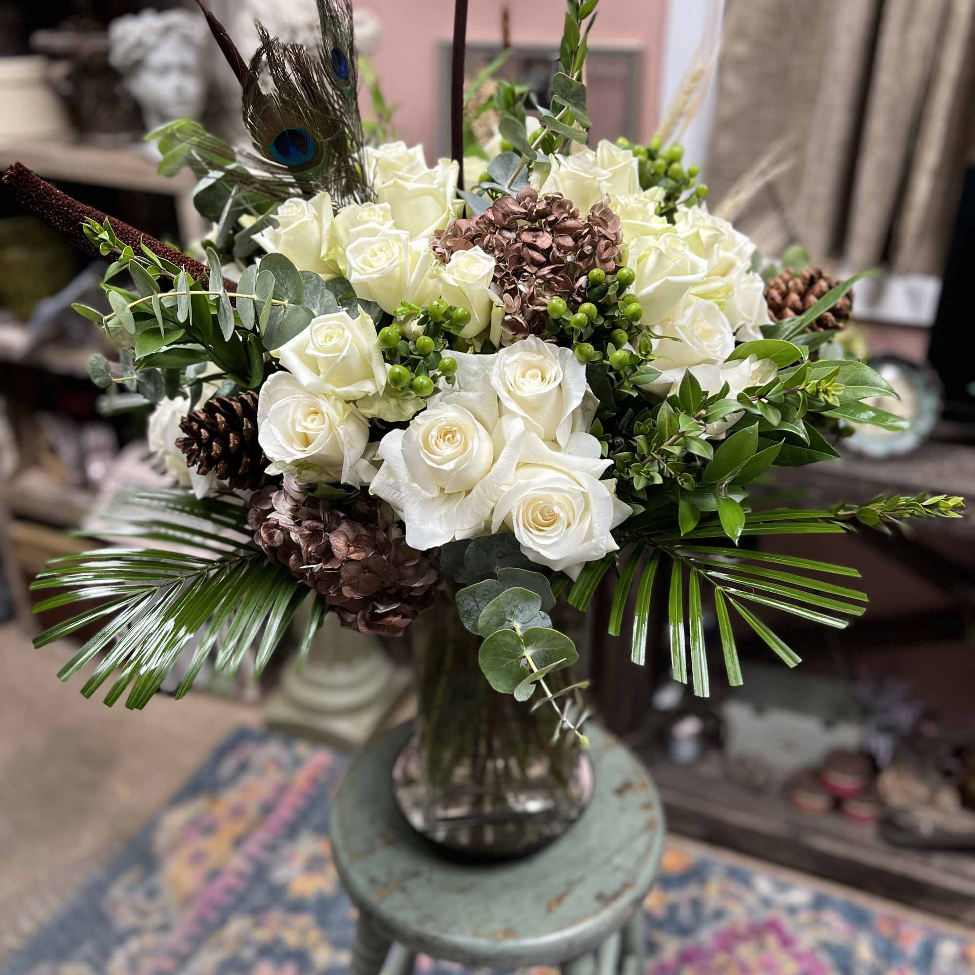 White roses arranged in a glass vase with peacock feathers and pinecones