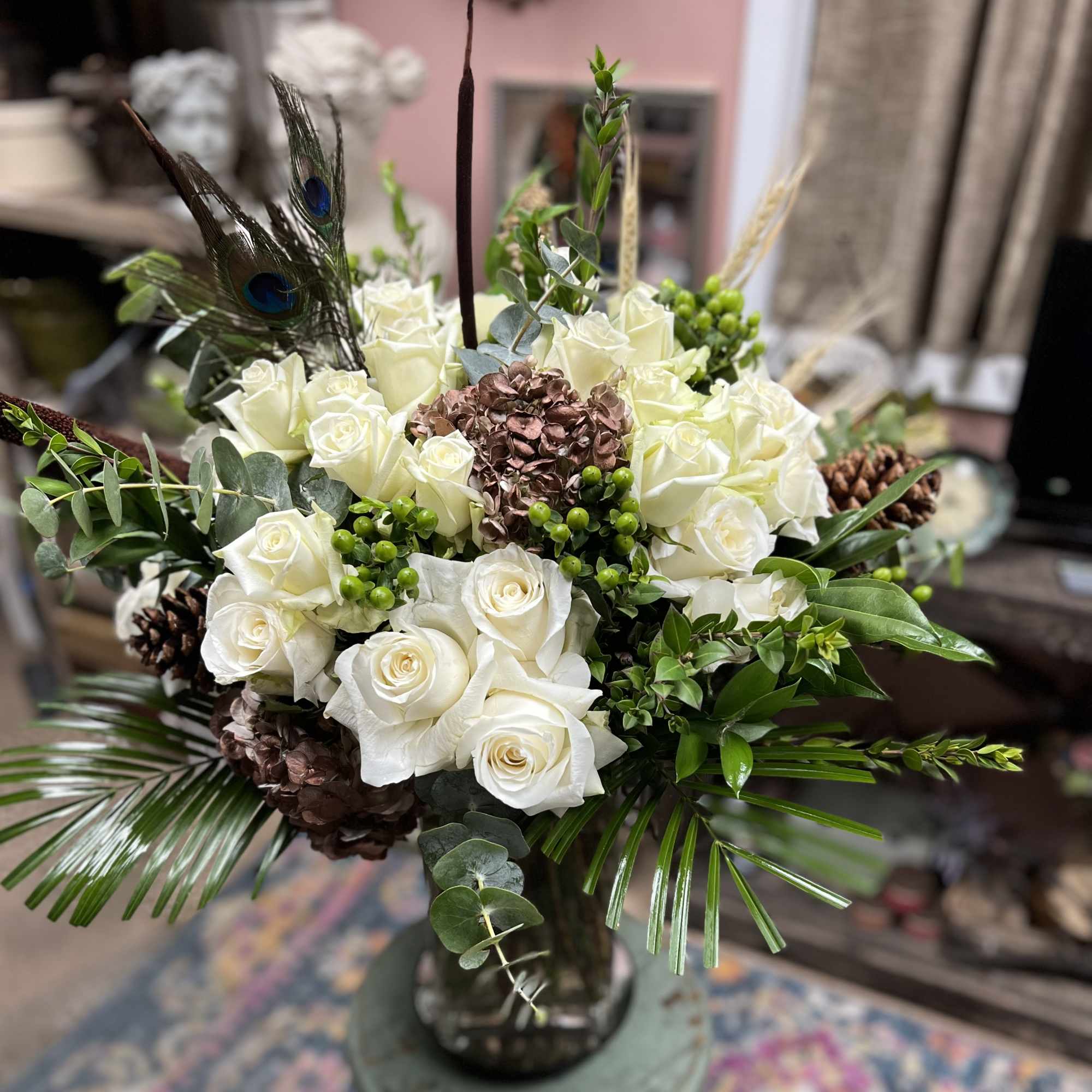 White rose arrangement with peacock feathers and pinecones in a glass vase