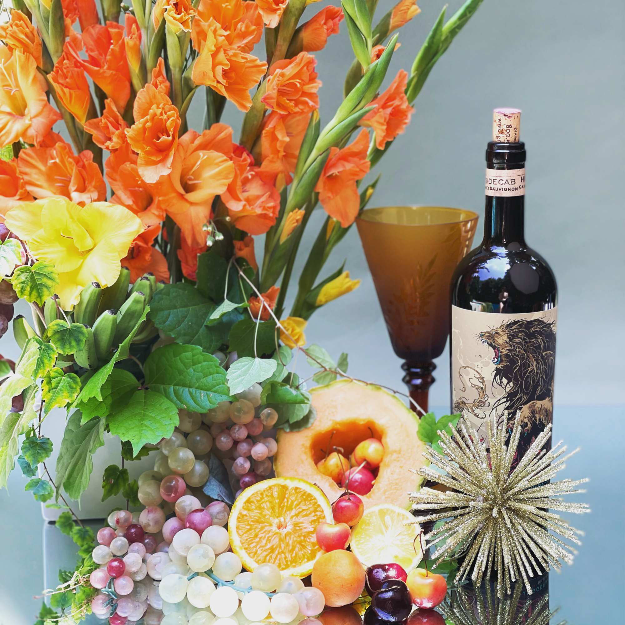 Orange gladiolus arrangement with fruit, grapes, and a wine bottle