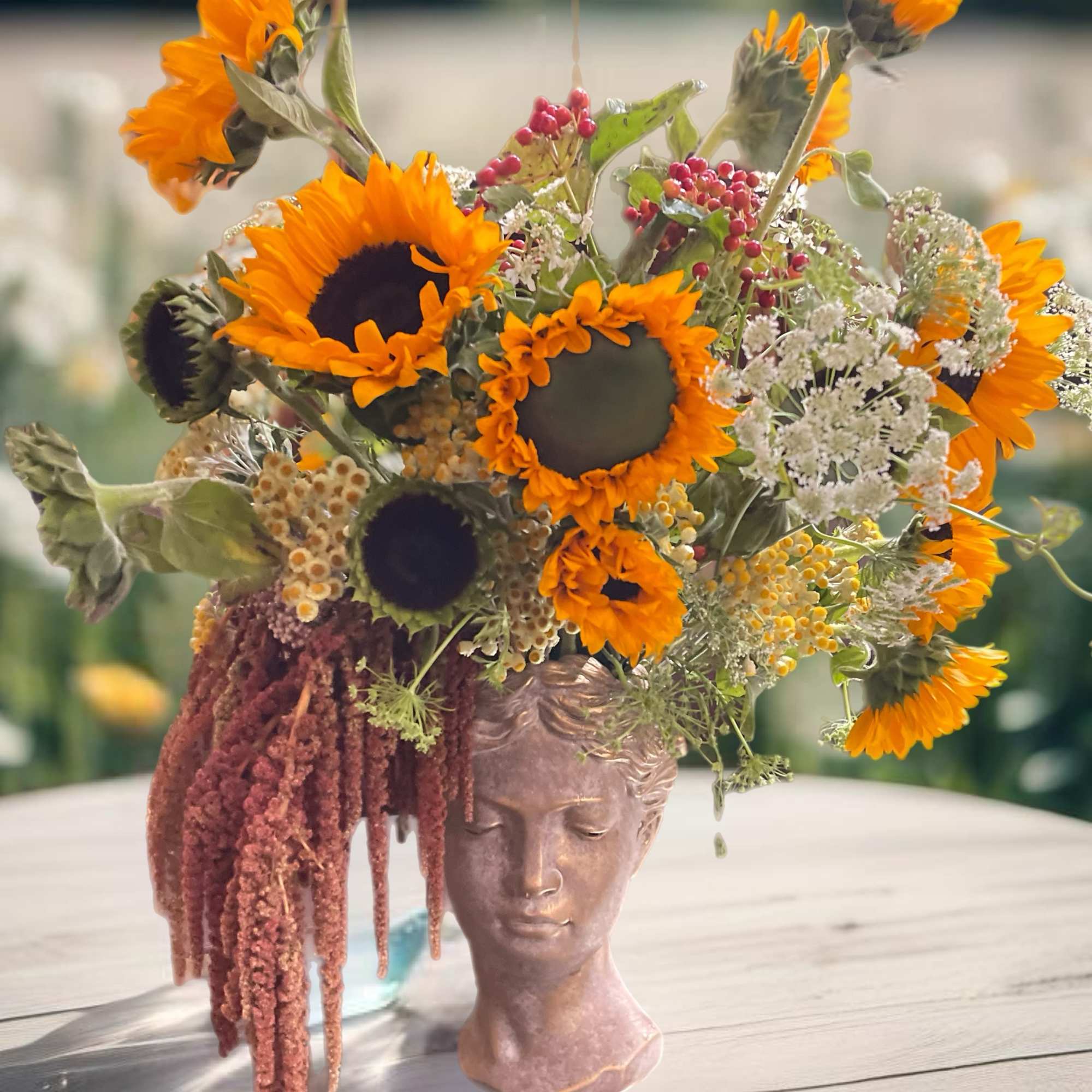 Sunflower arrangement in a sculpted face vase with trailing burgundy amaranthus